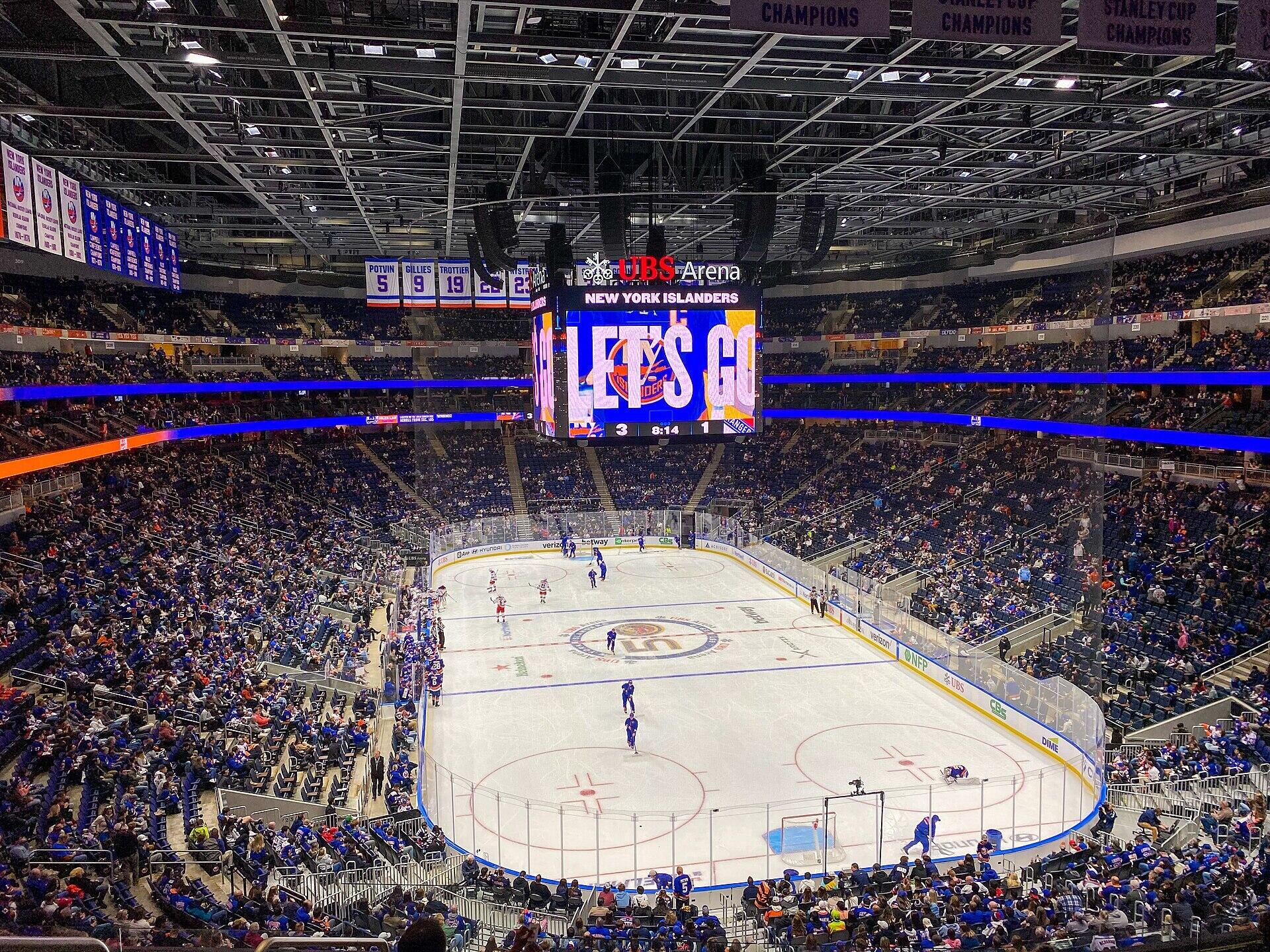 A vibrant hockey game at UBS Arena, featuring the New York Islanders with a packed crowd and a large scoreboard displaying "LET'S GO."