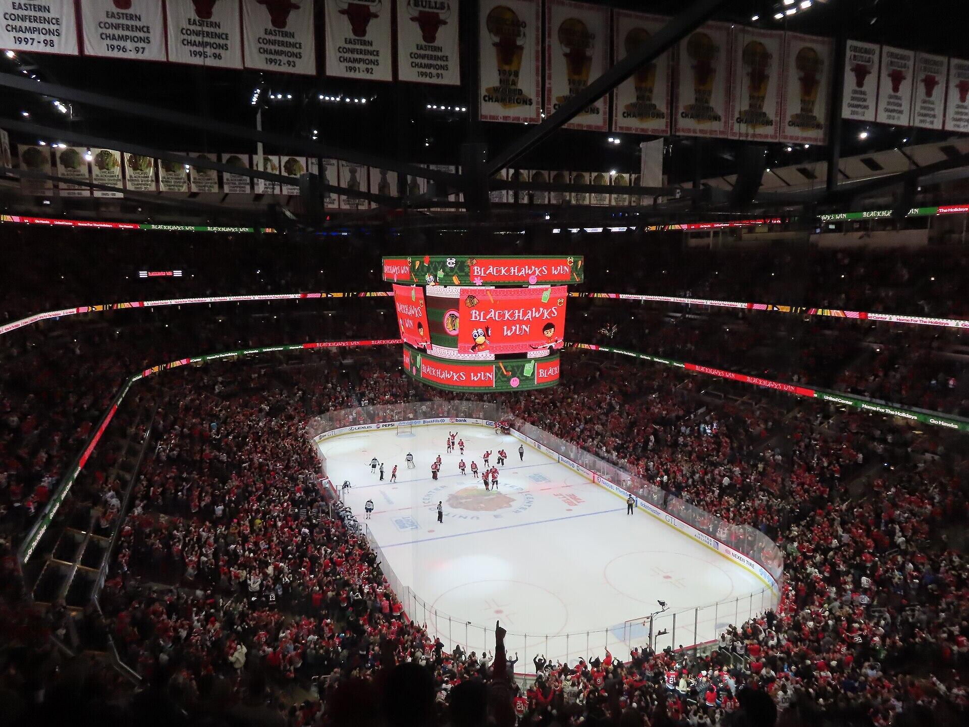 Crowd cheers in a packed arena as the scoreboard announces a Chicago Blackhawks victory in a hockey game.