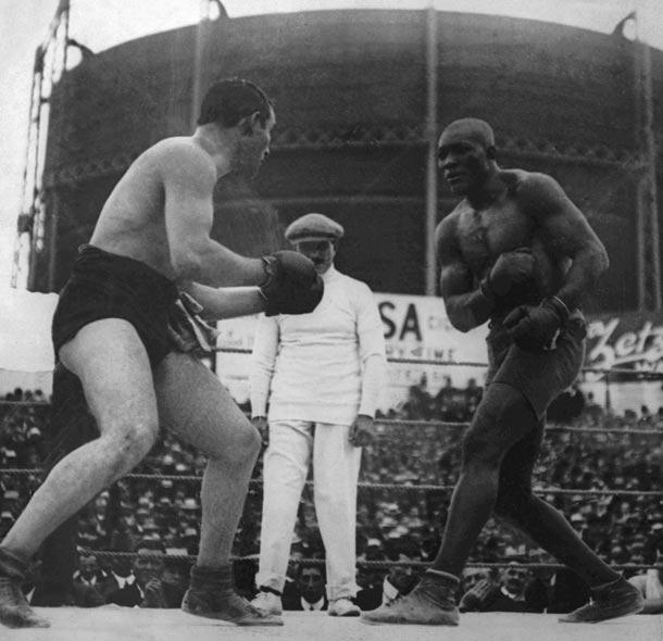 Black and white photo of two boxers facing off in a ring with a referee standing between them and a large crowd watching.