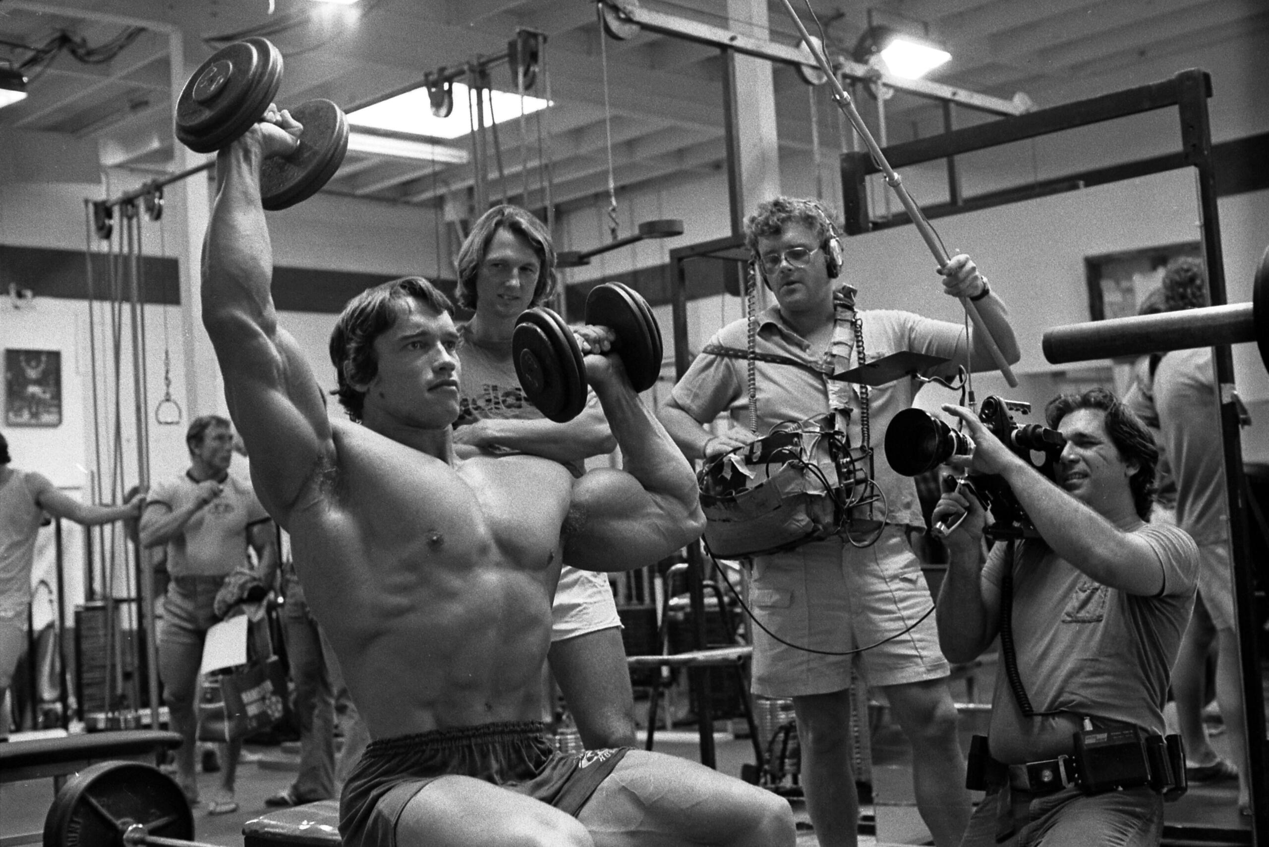 Black-and-white photo of a muscular man lifting dumbbells in a gym while a film crew records him.