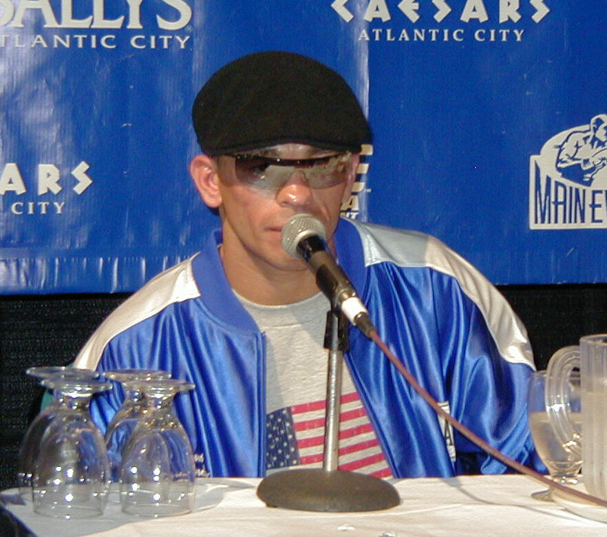 Person wearing a blue jacket and American flag shirt speaking into a microphone at a press event with Atlantic City casino logos in the background.