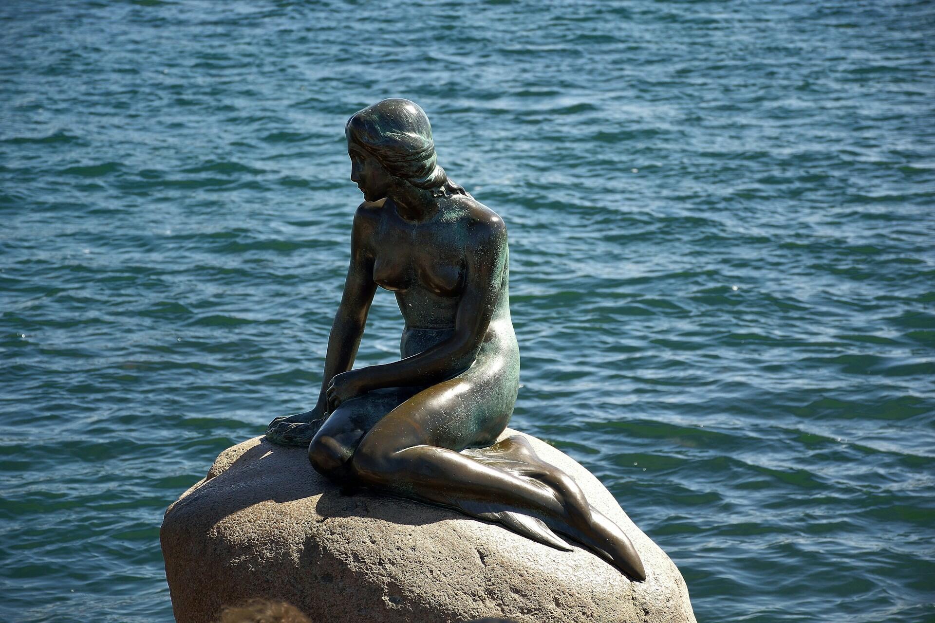 Statue of a seated mermaid on a rock surrounded by water, with a blurred face.