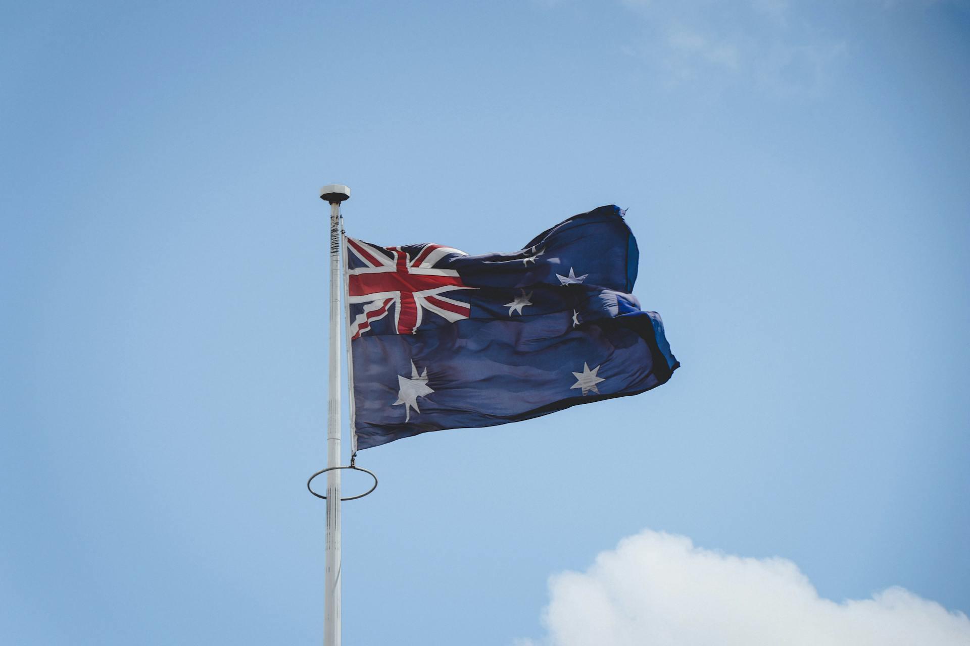 Australian flag waving on a flagpole against a clear blue sky with a small white cloud.
