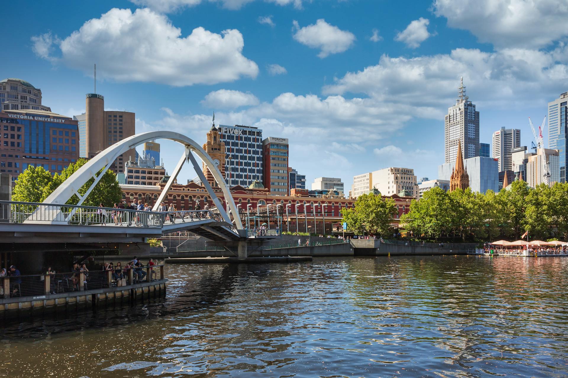 A river flows beneath a bridge, with buildings lining the background against a clear sky.
