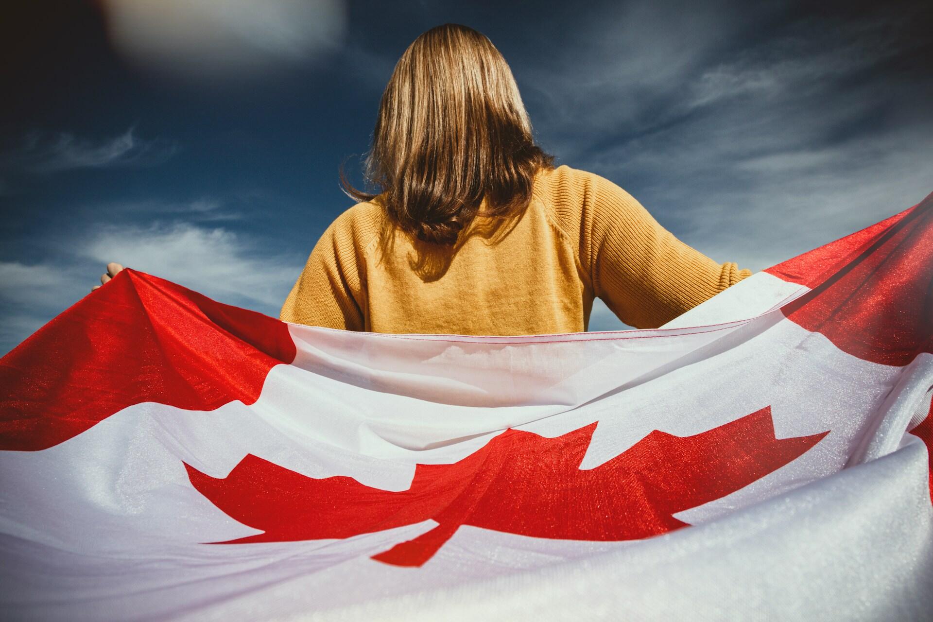 Person wearing a mustard sweater holding a large Canadian flag against a partly cloudy blue sky background.