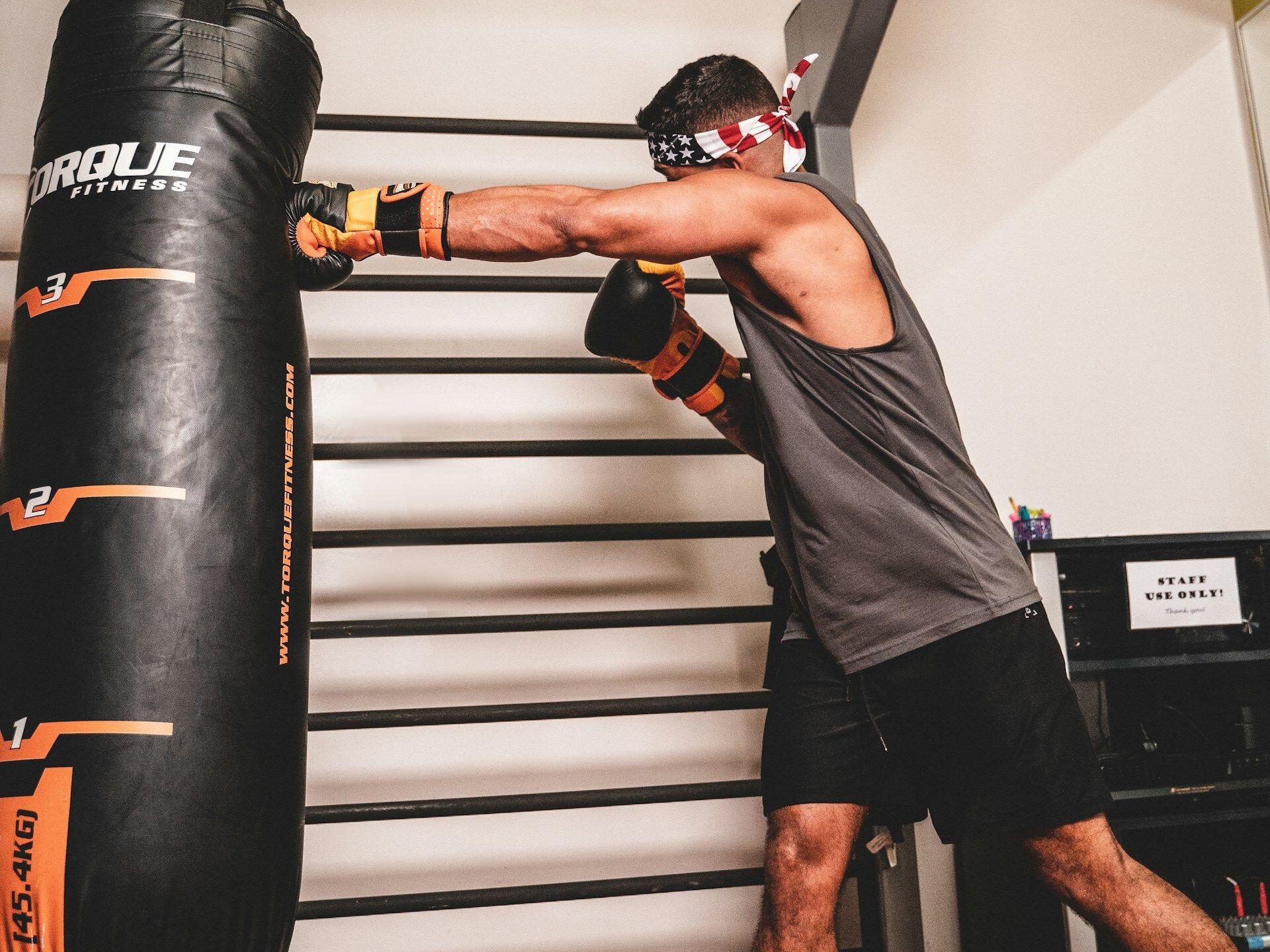 Man wearing an American flag bandana and boxing gloves practices punching a heavy bag in a gym setting.