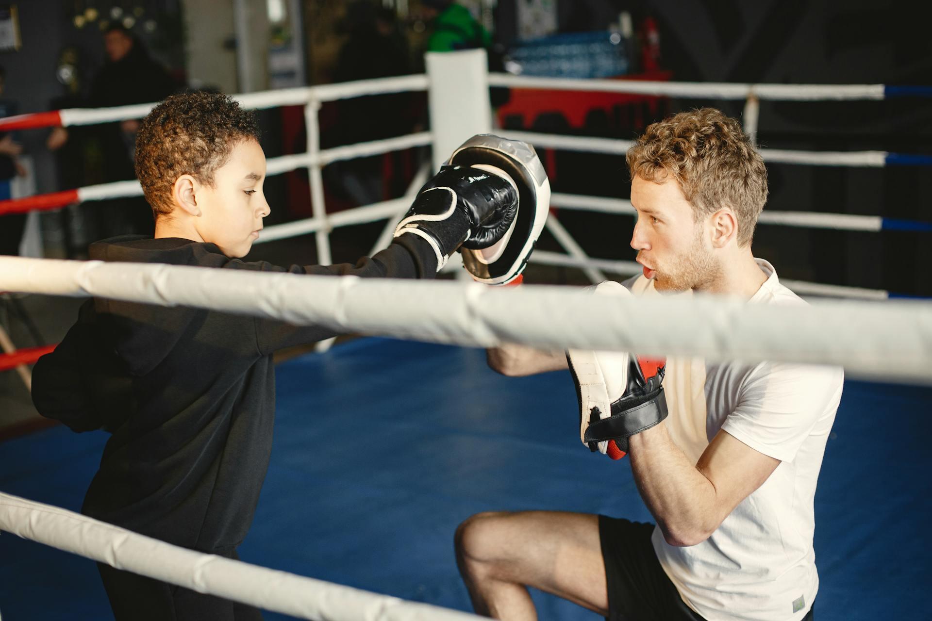 A young boxer practices techniques with a coach in a boxing ring, wearing protective gloves and focused on training.