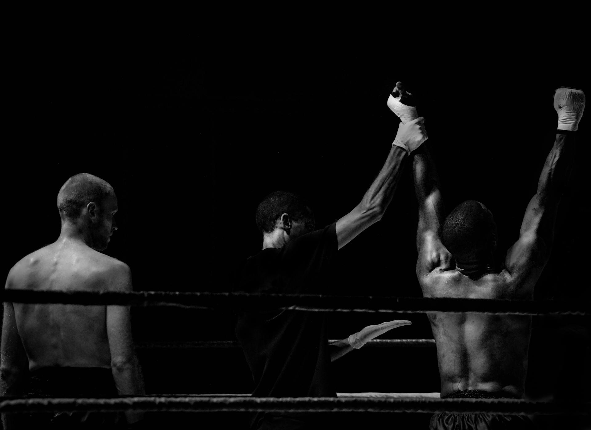 Boxers celebrate a victory in a dimly lit ring, with one fighter's arm raised by a referee amidst a dramatic black backdrop.