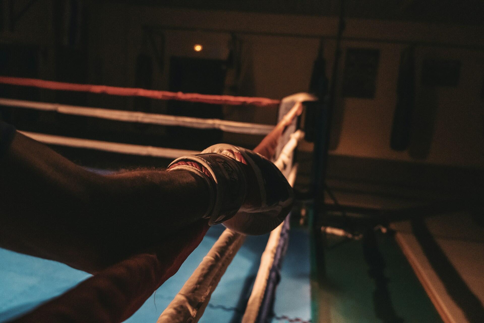 A close-up of a gloved hand resting on a boxing ring's red and white ropes, illuminated in a dimly lit gym.