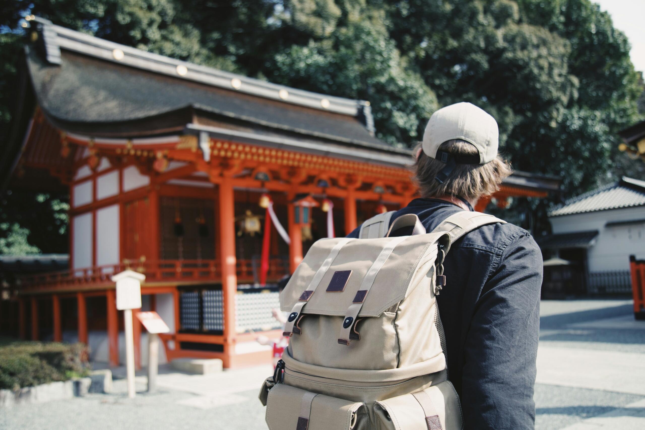 Person with beige backpack and cap standing near traditional Japanese shrine with orange pillars and black roof.
