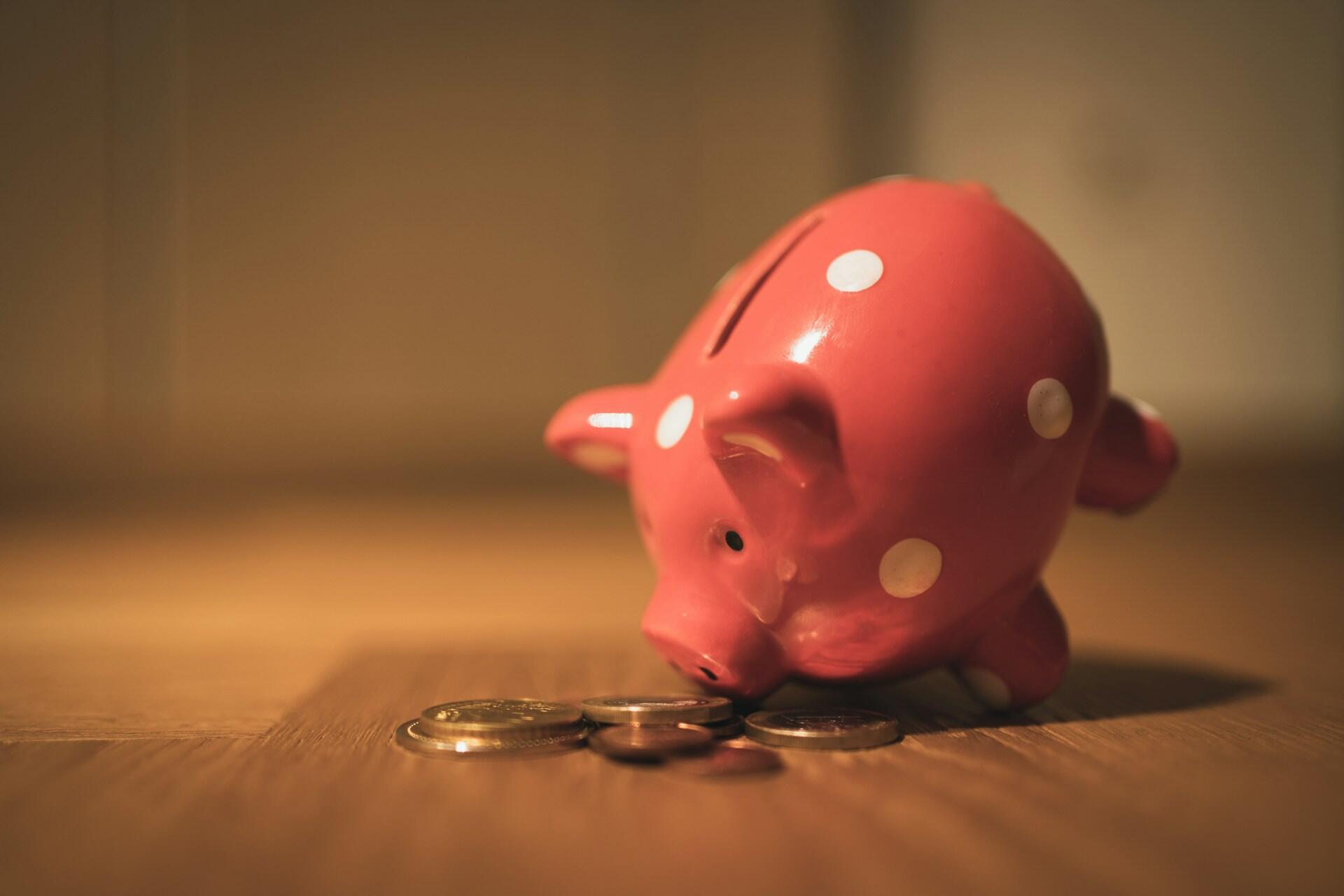 Pink piggy bank with white spots tipped over on wooden floor next to scattered coins.