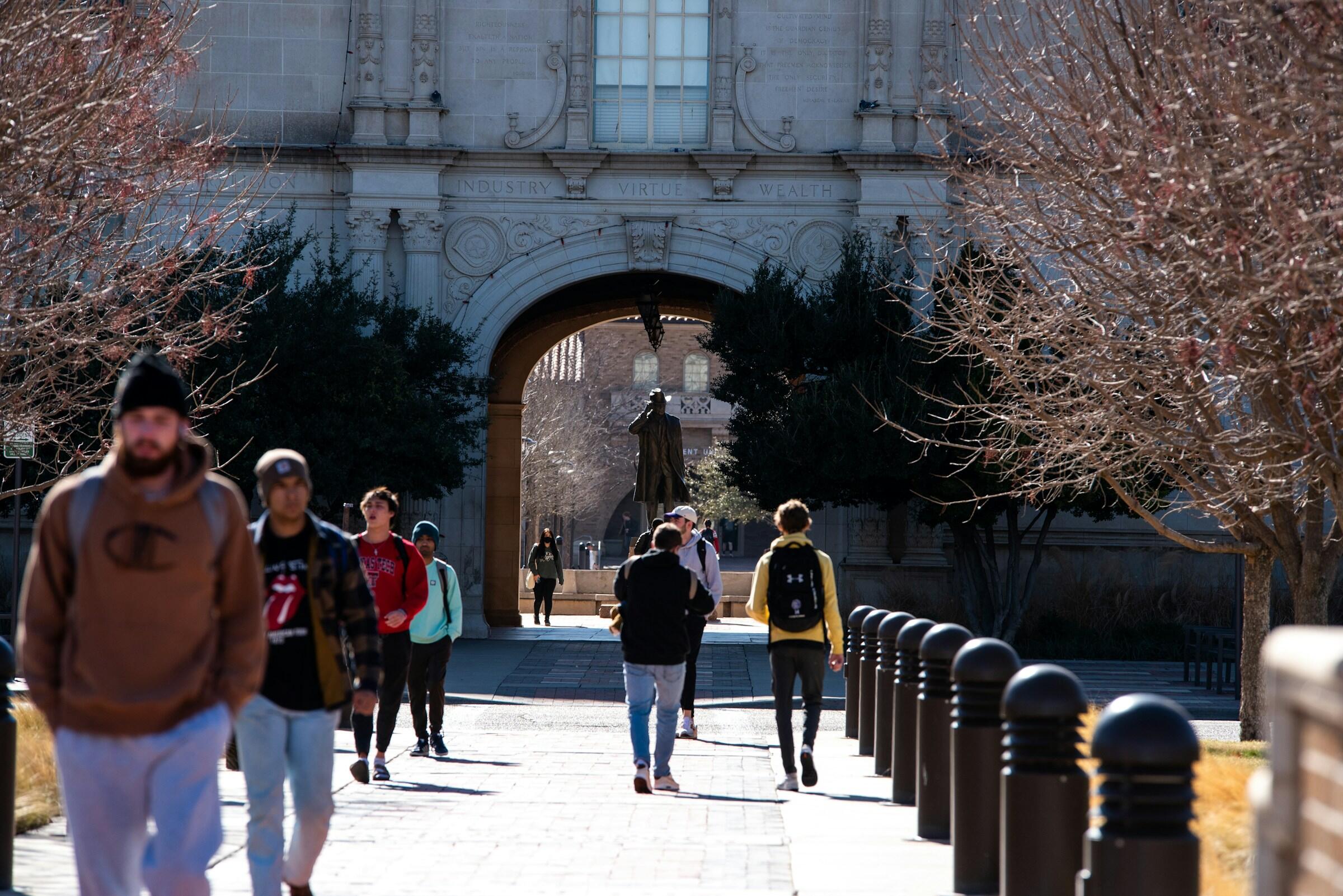 Students walk along a tree-lined pathway under an archway with the words "Industry, Virtue, Wealth" inscribed above.