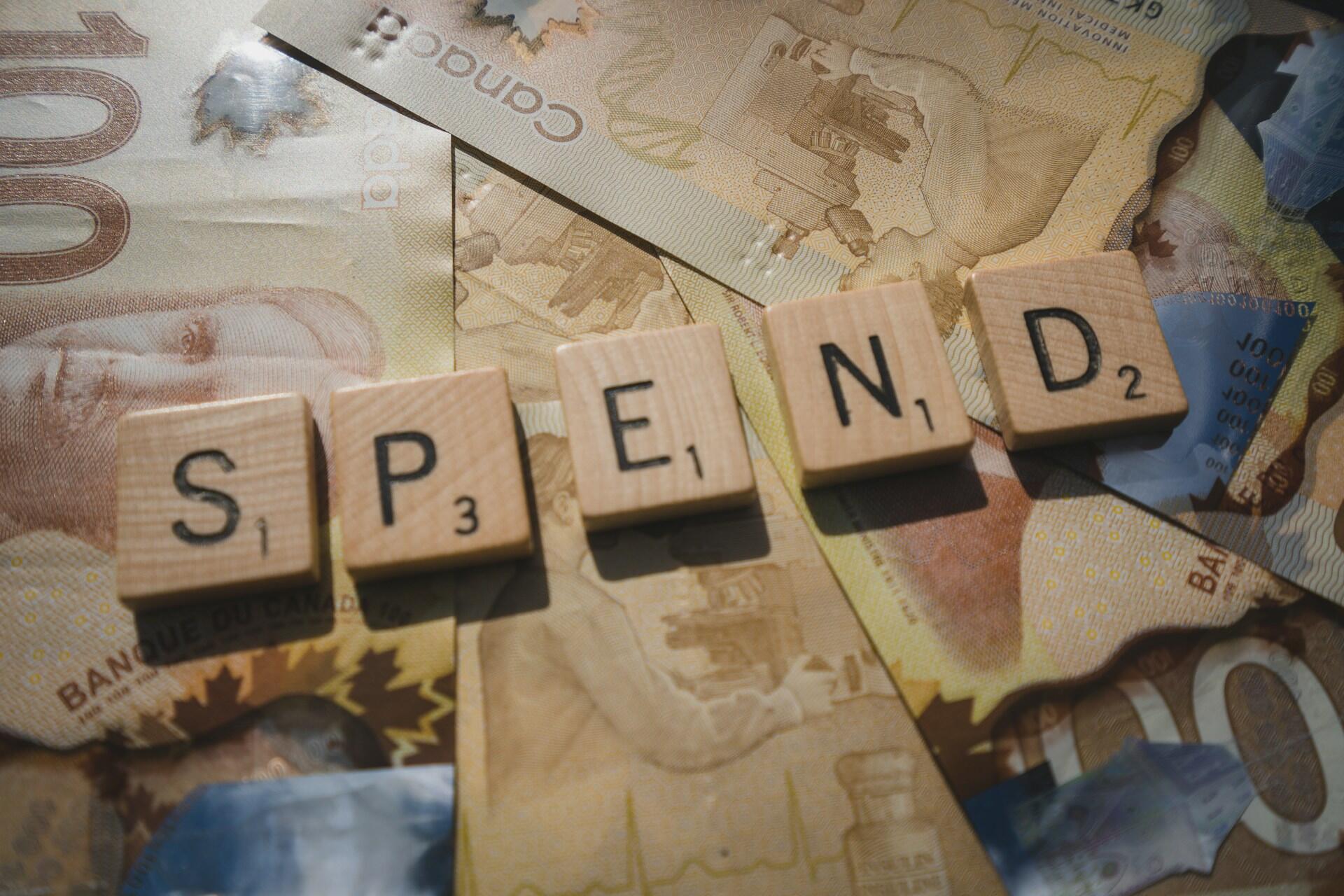 Wooden scrabble tiles spelling "SPEND" on top of Canadian 100-dollar bills.