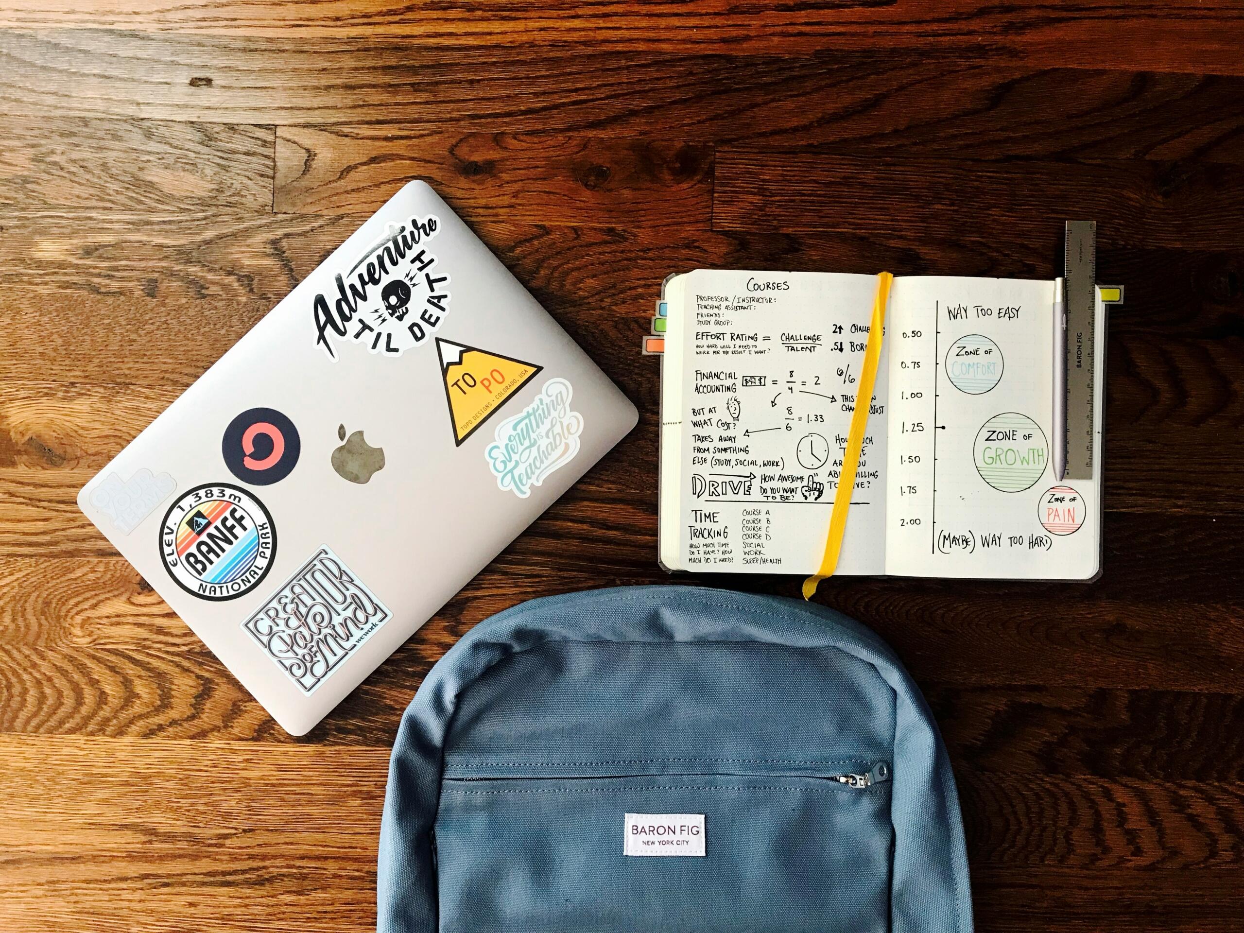 Silver laptop with stickers, an open notebook with handwritten notes and diagrams, and a blue backpack on a wooden floor.