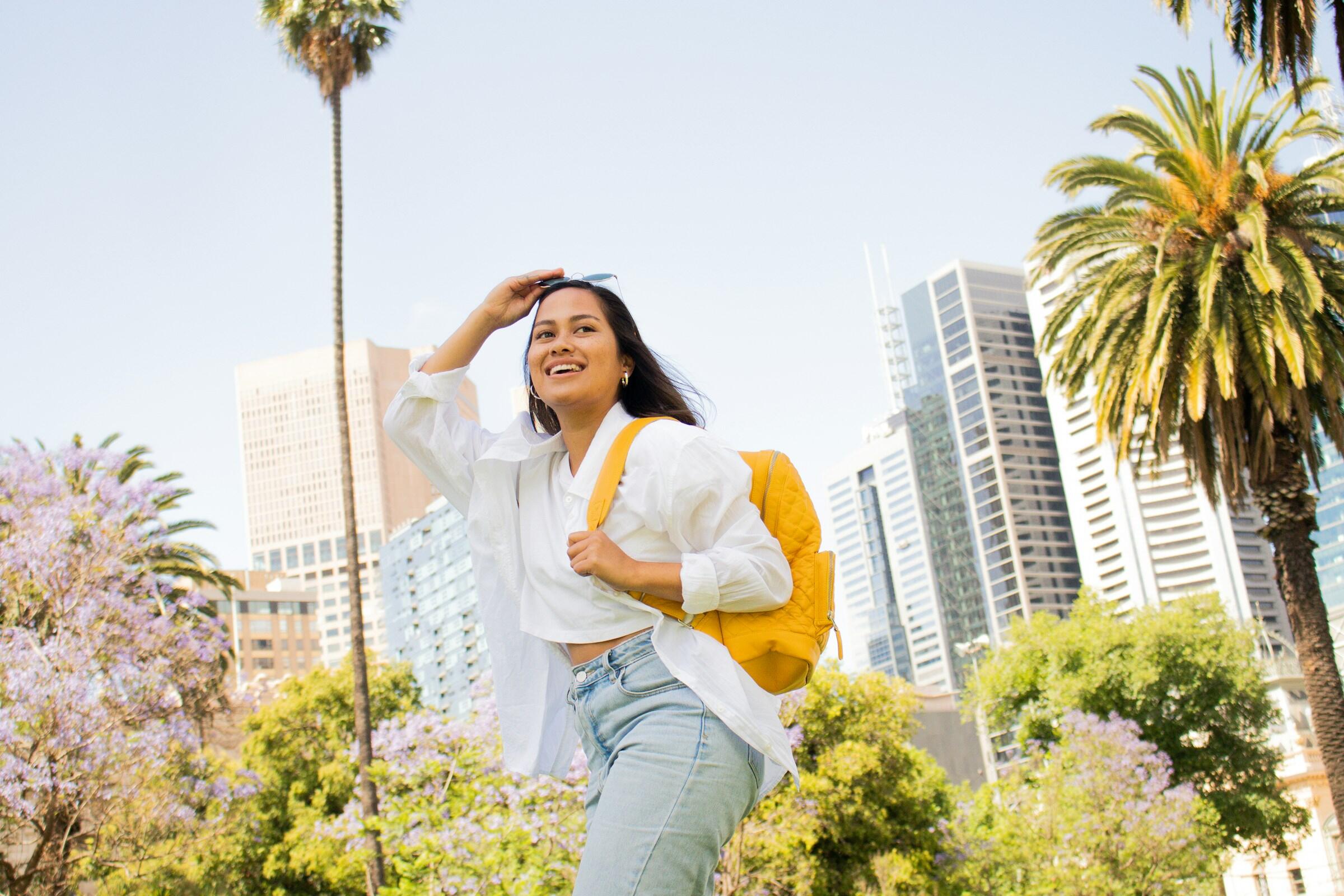 A woman in jeans and a white shirt walks through a park, surrounded by greenery and trees.