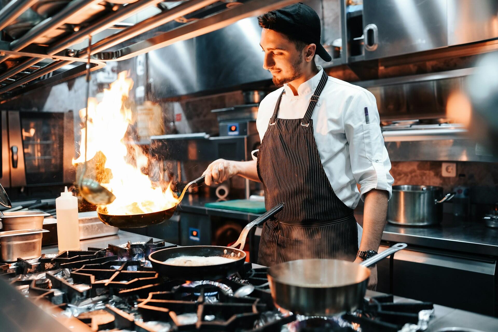 A chef skillfully tosses food in a flaming pan in a busy restaurant kitchen, showcasing culinary expertise and vibrant kitchen activity.