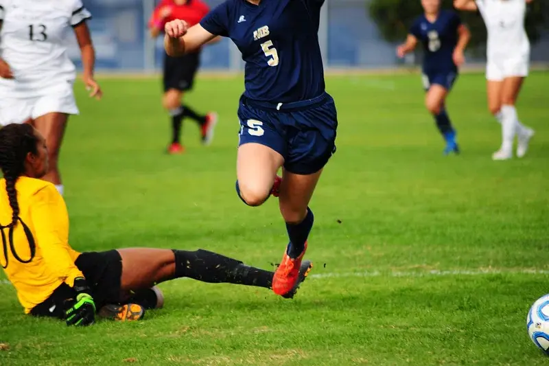 Soccer player in navy jersey evades sliding goalkeeper in yellow against players in white on green field during a match.