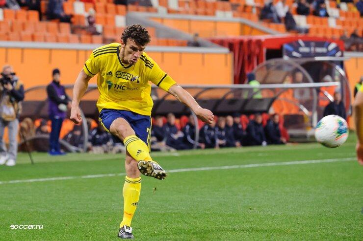 Soccer player in yellow uniform kicks the ball during a match on a grass field with spectators in the background.