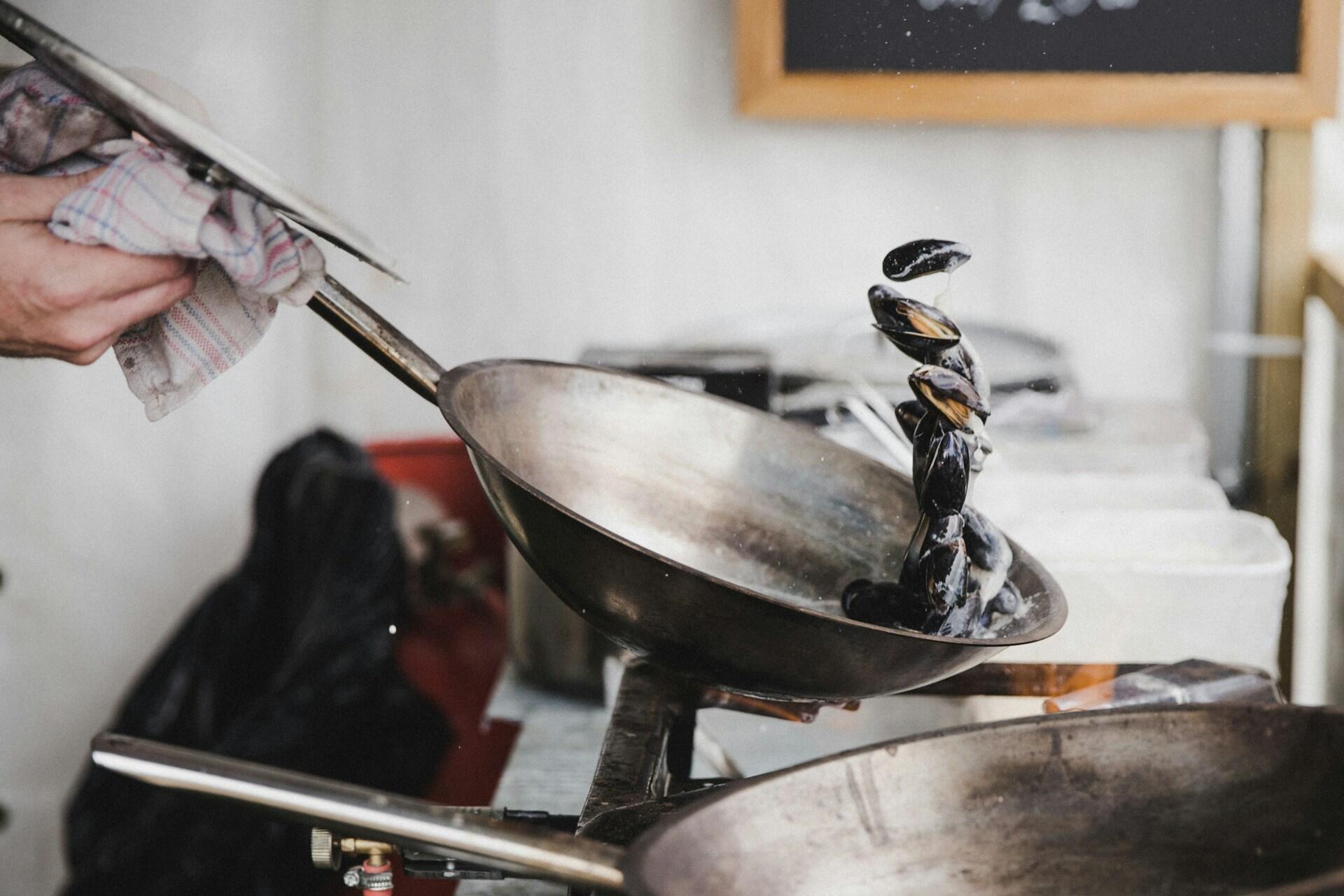 A chef lifts a frying pan with mussels, water splashing as the mussels swirl above another pan in a bustling kitchen.
