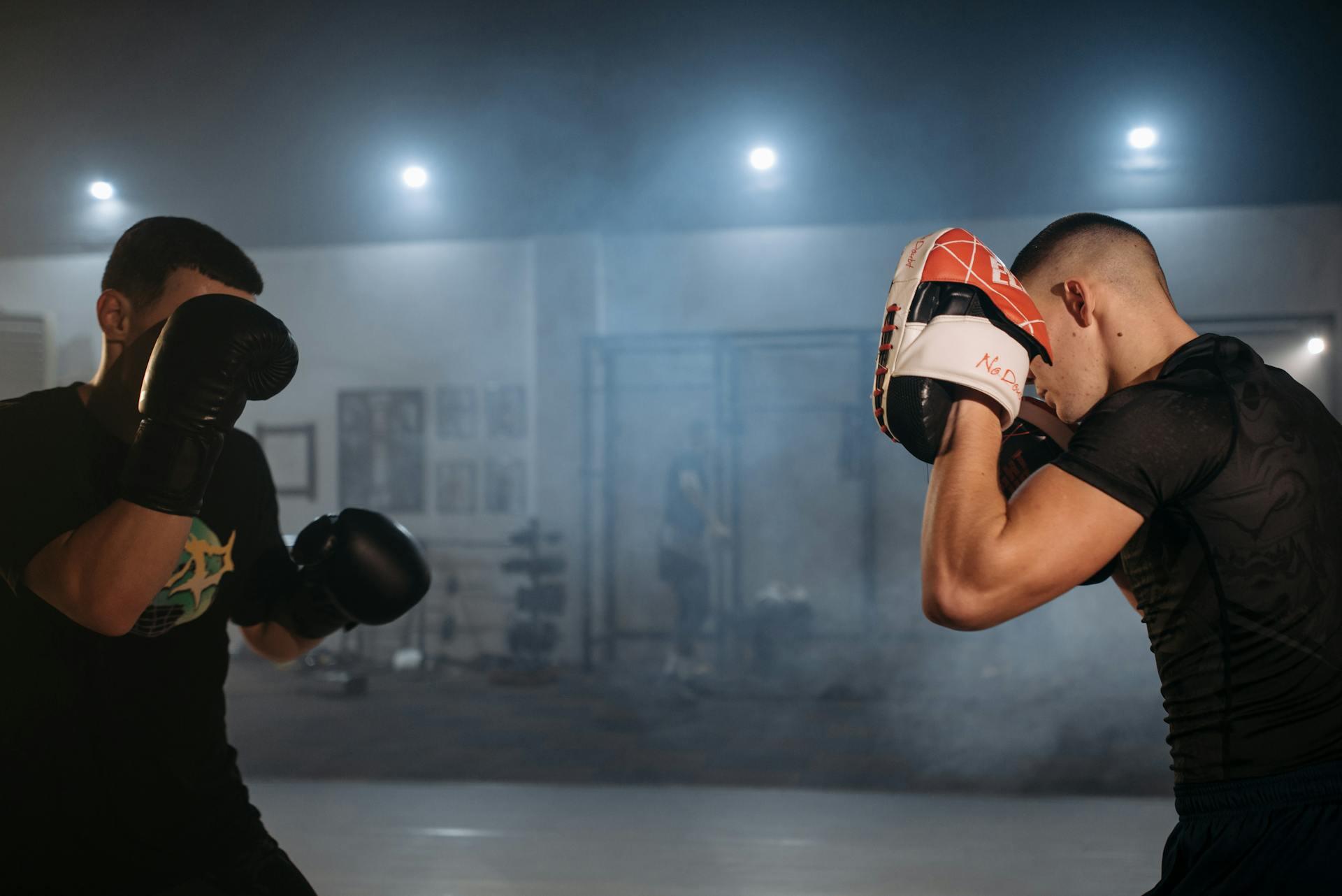 Two male fighters practice boxing techniques in a dimly lit gym filled with light smoke, showcasing their focus and stance.