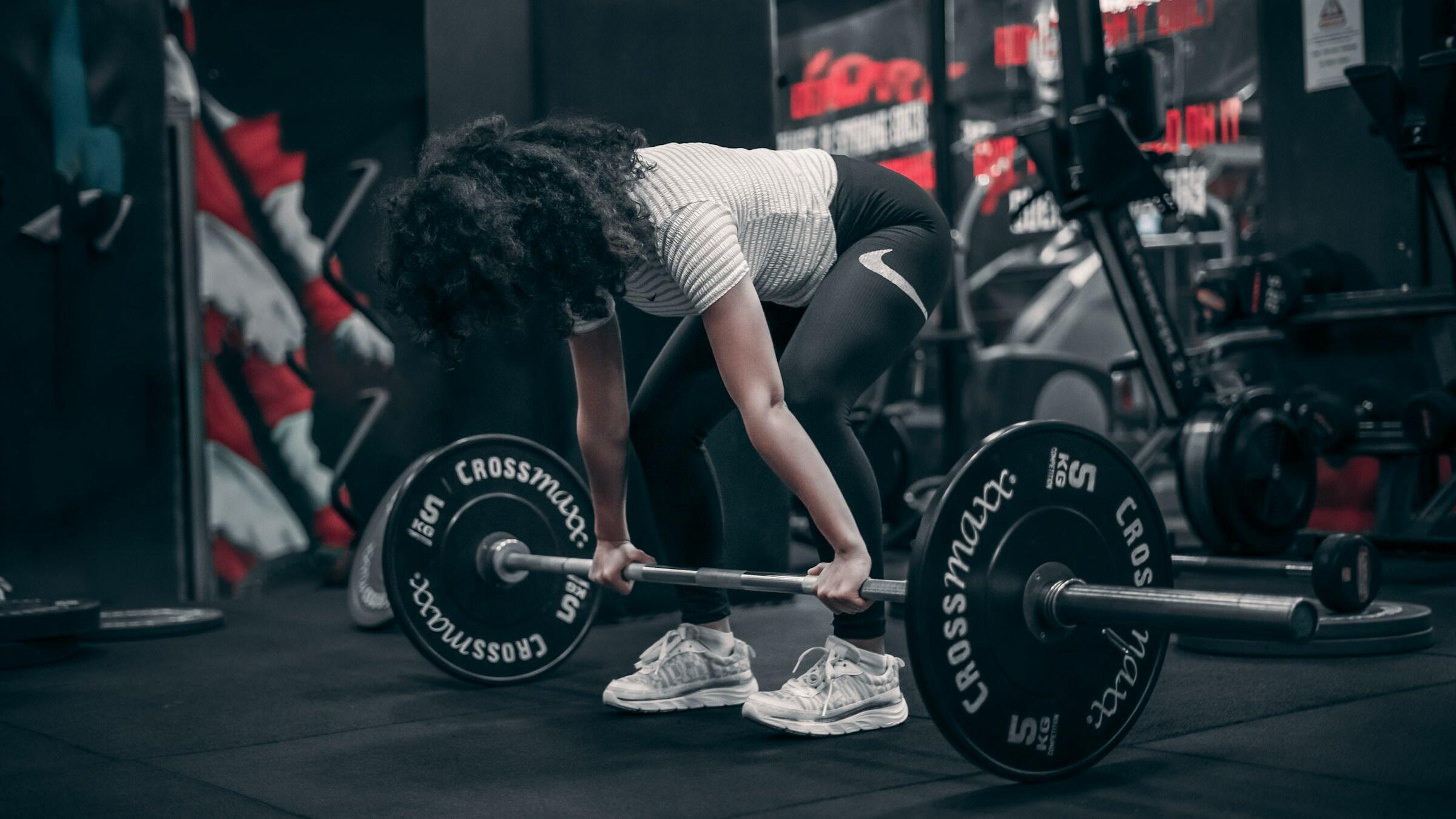 Woman with curly hair lifting a barbell in a gym, wearing a striped shirt, black leggings, and white sneakers.