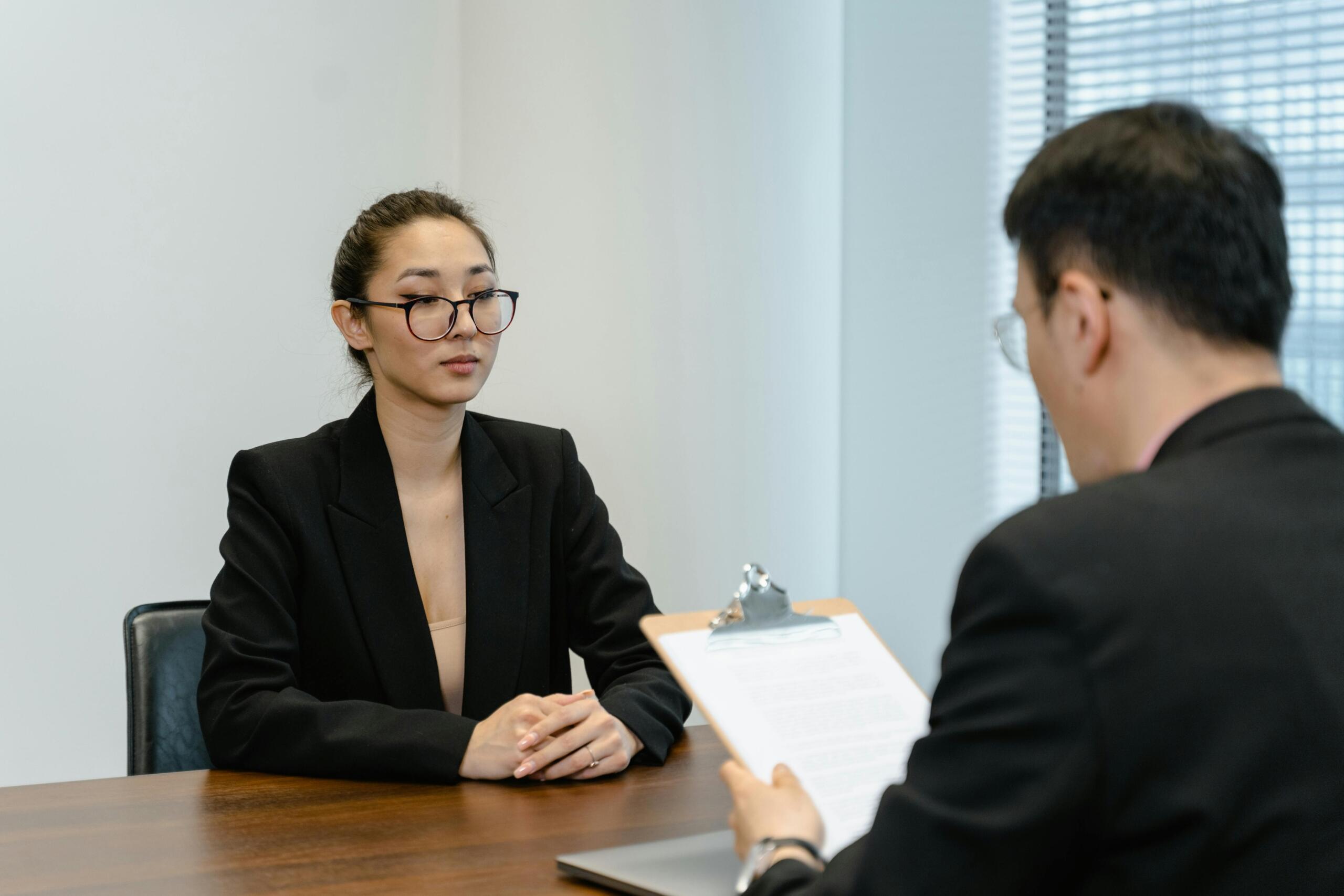 Woman sitting at a table during a job interview, speaking with an interviewer across from her in a professional office setting.