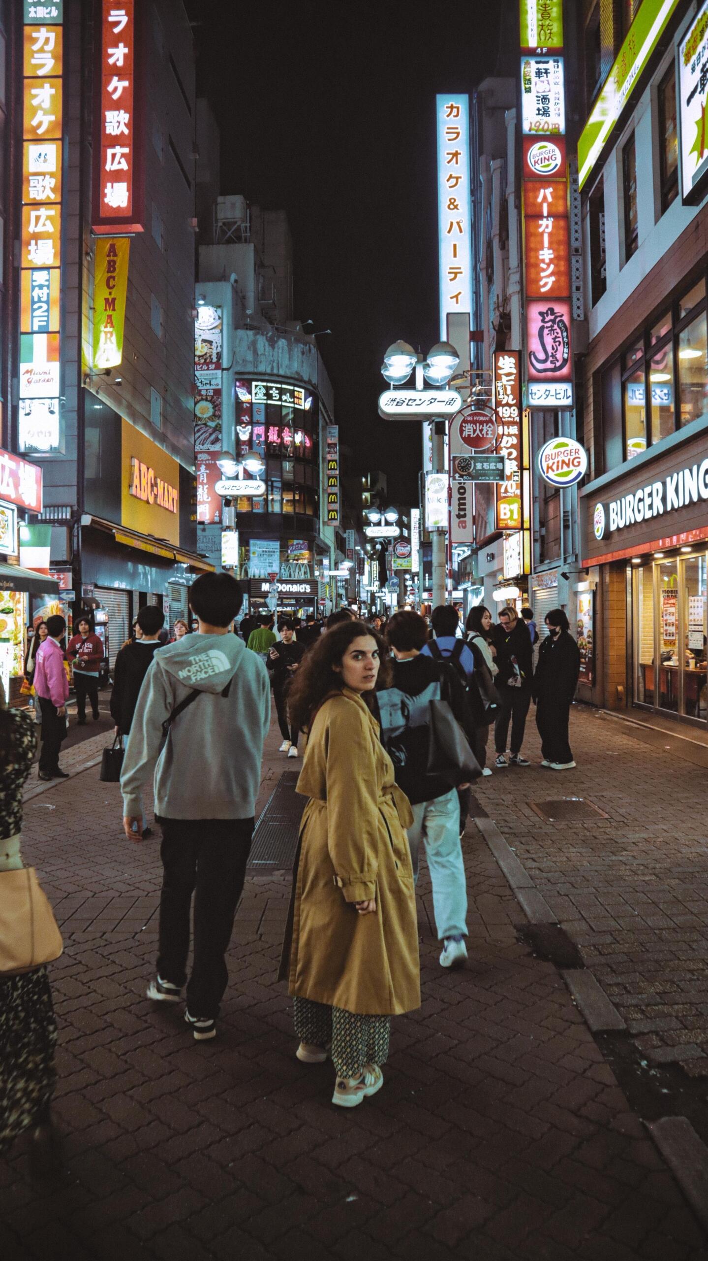 Foreigner standing on a brightly lit street in Japan at night, surrounded by neon signs, storefront lights, and a busy urban atmosphere.
