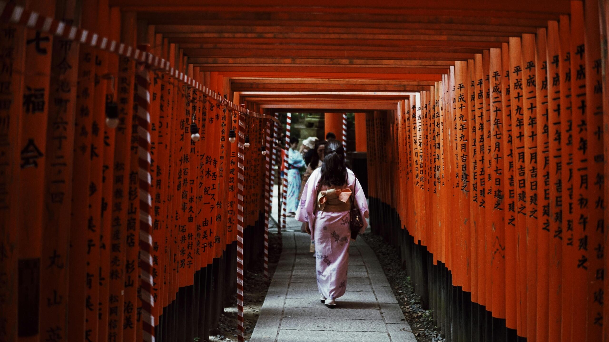Japanese woman walking through a famous shrine pathway lined with bright orange torii gates, with the repeating arches creating a traditional and scenic atmosphere.
