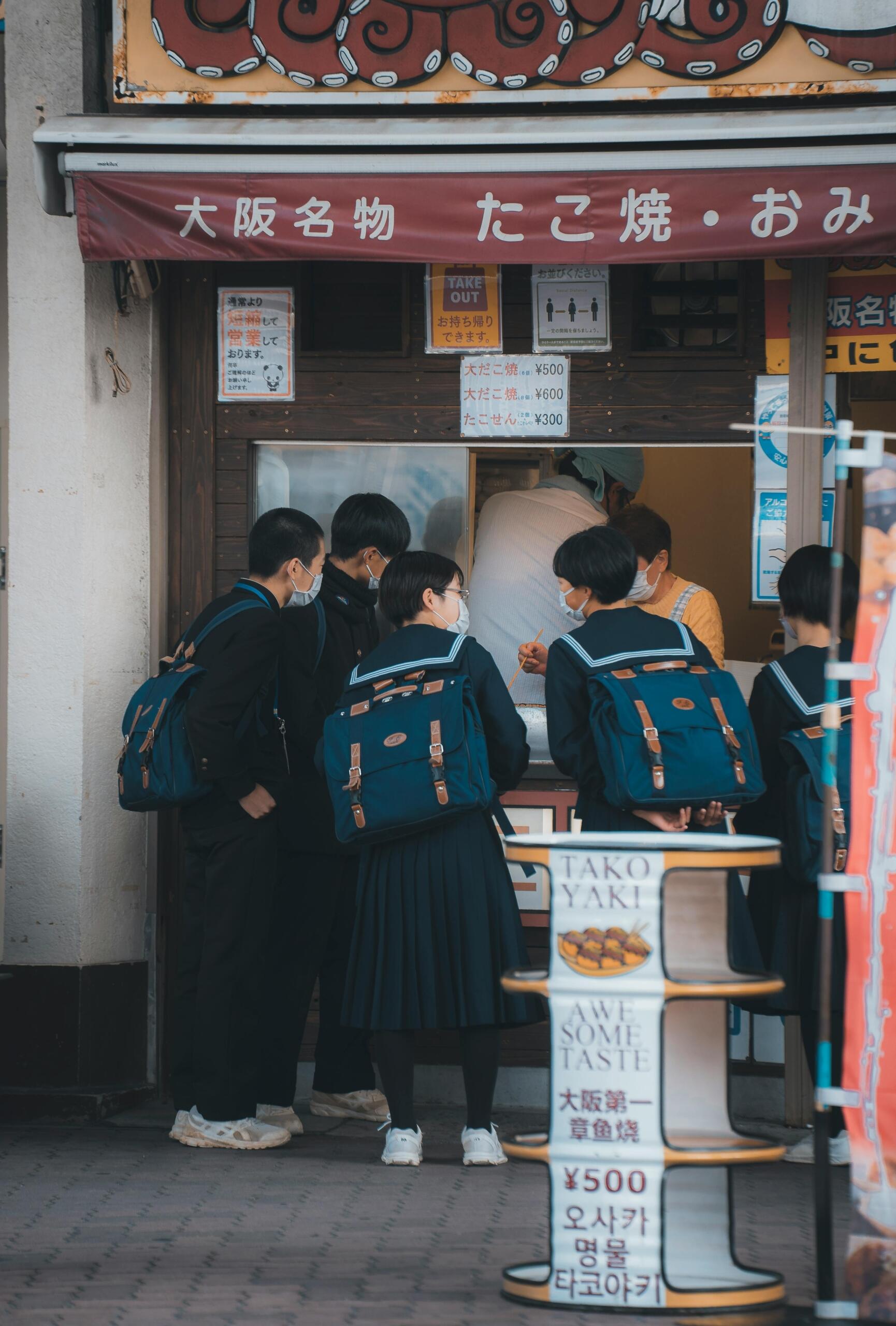 Group of Japanese students standing together inside a casual takeout restaurant, with food counters and menu signs visible in the background.