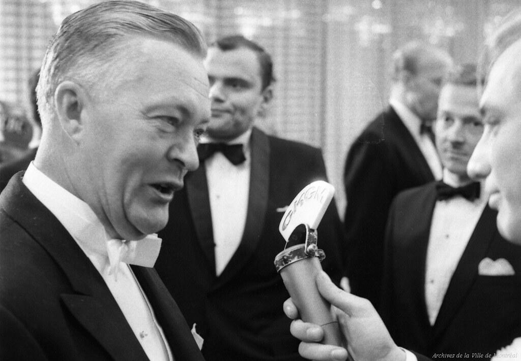 Black and white photo of men in tuxedos being interviewed with a microphone labeled "CHGM" at a formal event.