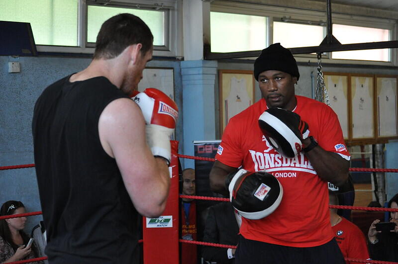 Two boxers sparring in a gym, one wearing red training gloves and a matching shirt, the other in a black sleeveless top.