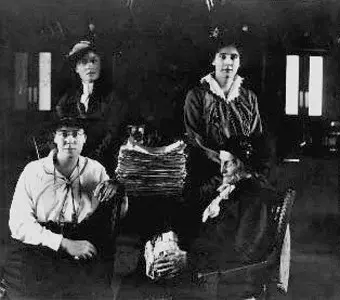 Black and white photo of four people, three seated and one standing, with stacks of papers on a table in a dimly lit room.