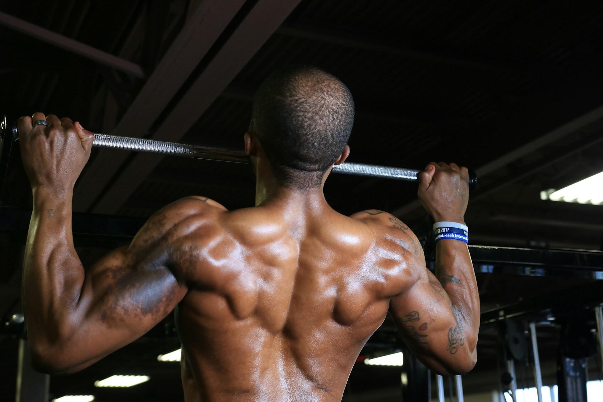 A muscular person performs pull-ups, showcasing defined back muscles and arms, in a gym environment with industrial lighting.