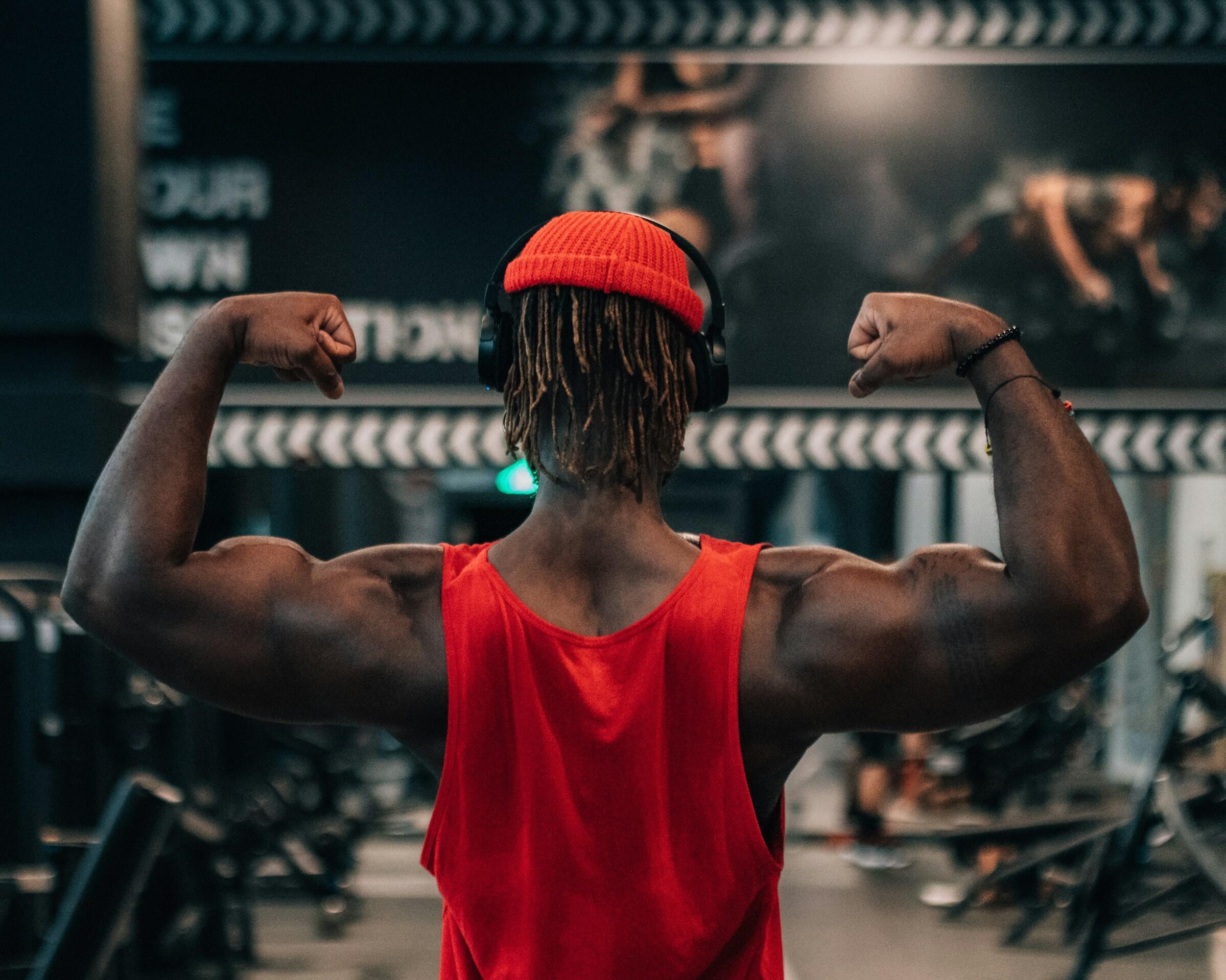 Muscular person wearing a red tank top and beanie flexes biceps facing a gym mirror while wearing headphones.