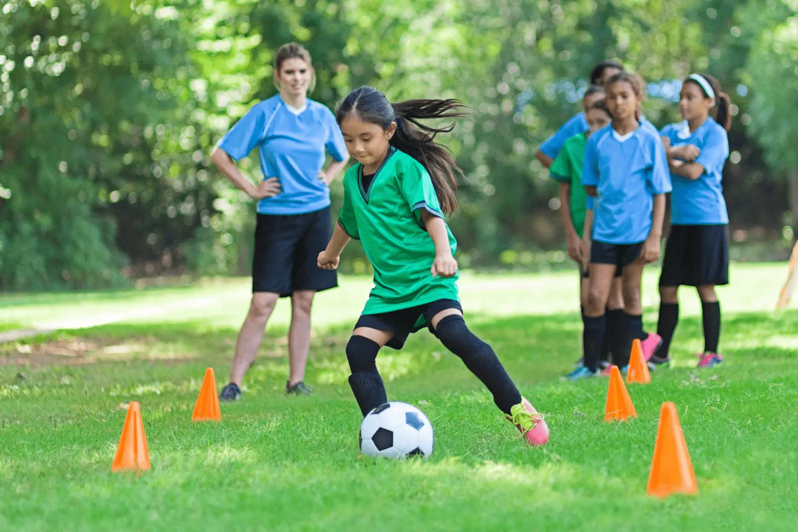Child in green jersey dribbling a soccer ball through cones while others in blue jerseys watch on a grassy field.