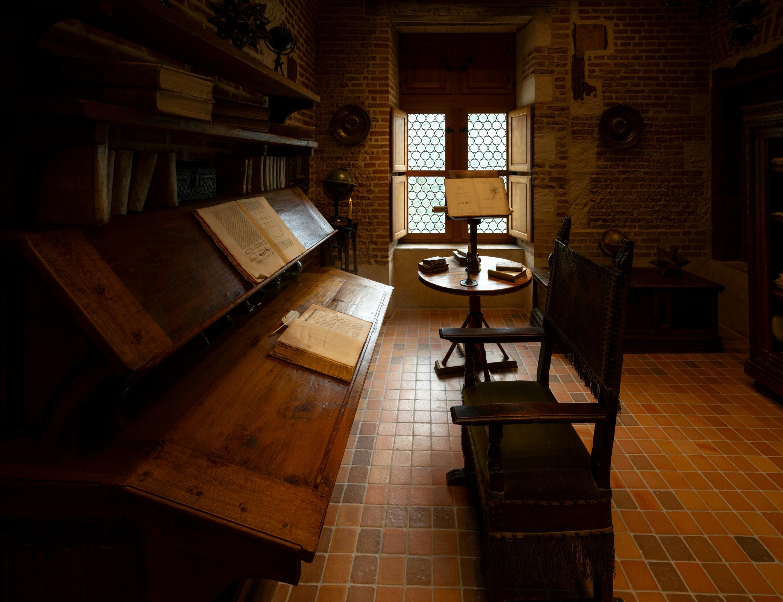 Cozy historic study room with wooden desks, antique books, a small round table, and a window with lattice panes letting in soft light.