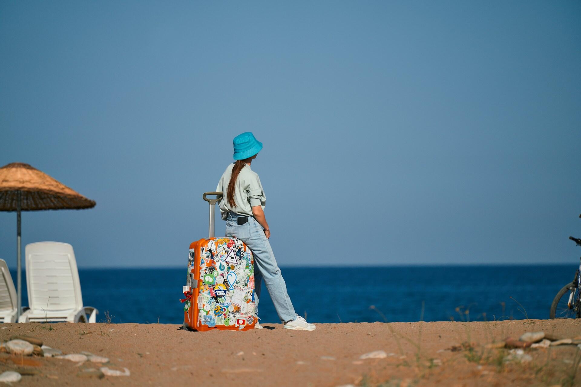 Person in blue bucket hat leaning on large orange suitcase covered with stickers on a sandy beach by the sea under clear blue sky.