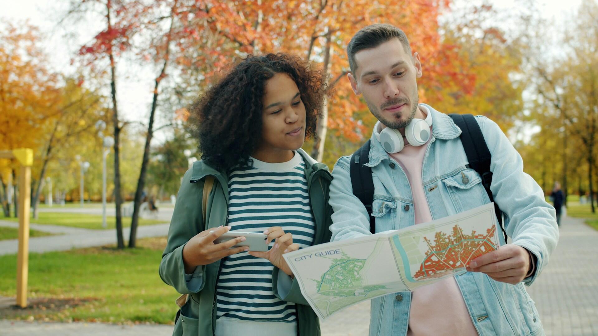 Two young adults outdoors in autumn, one holding a city guide map, the other using a smartphone, surrounded by colorful fall trees.