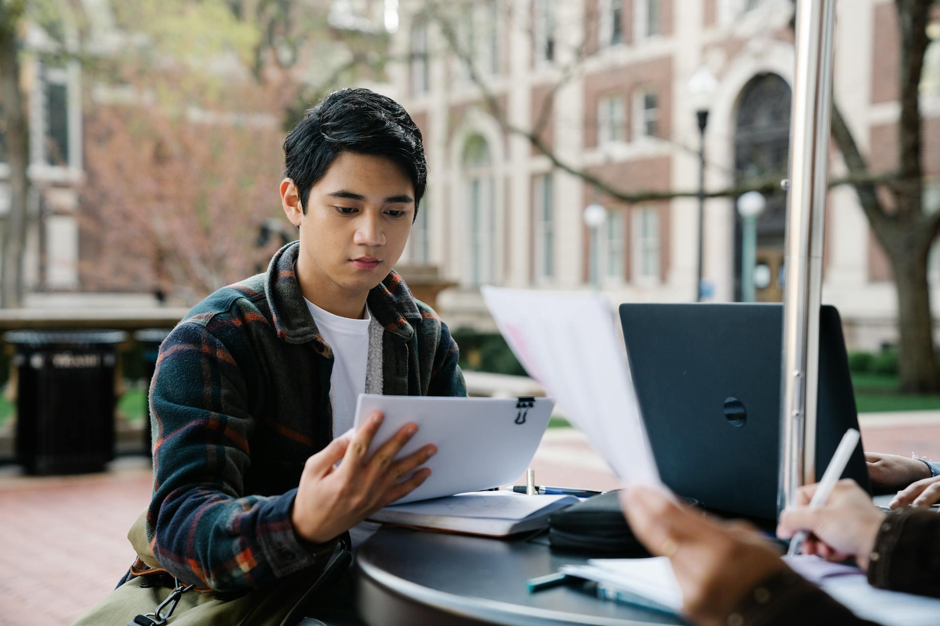 Two students studying outdoors at a table with papers and a laptop in a campus courtyard setting.