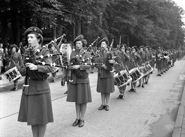 Black and white photo of a female military band marching and playing bagpipes and drums along a tree-lined street.