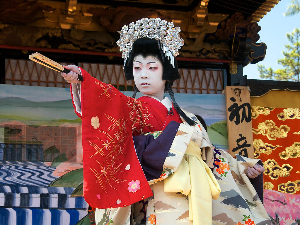 Performer in traditional Japanese costume and ornate headdress holding a fan on stage with painted backdrop and wooden sign.