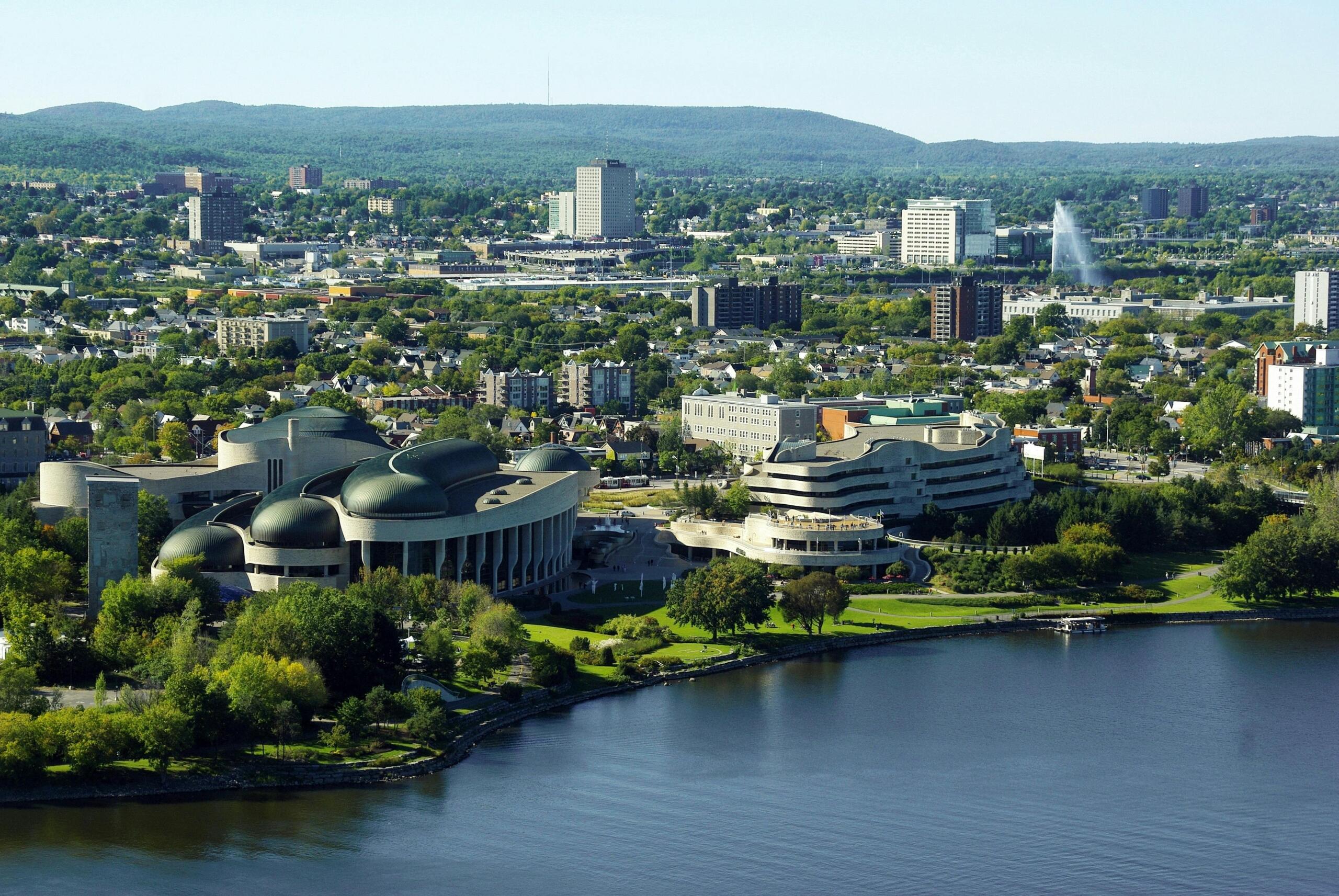 Aerial view of Ottawa showing the downtown skyline, river, bridges, historic buildings, and surrounding neighbourhoods under a clear sky.