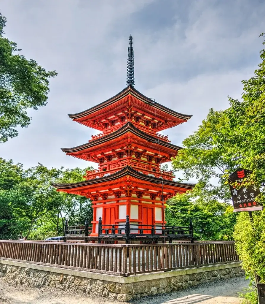 Bright red three-tiered Japanese pagoda surrounded by green trees under a partly cloudy sky.