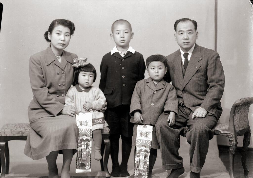 Black and white photo of a family with mother, father, and three children holding decorative paper banners, seated and standing indoors.