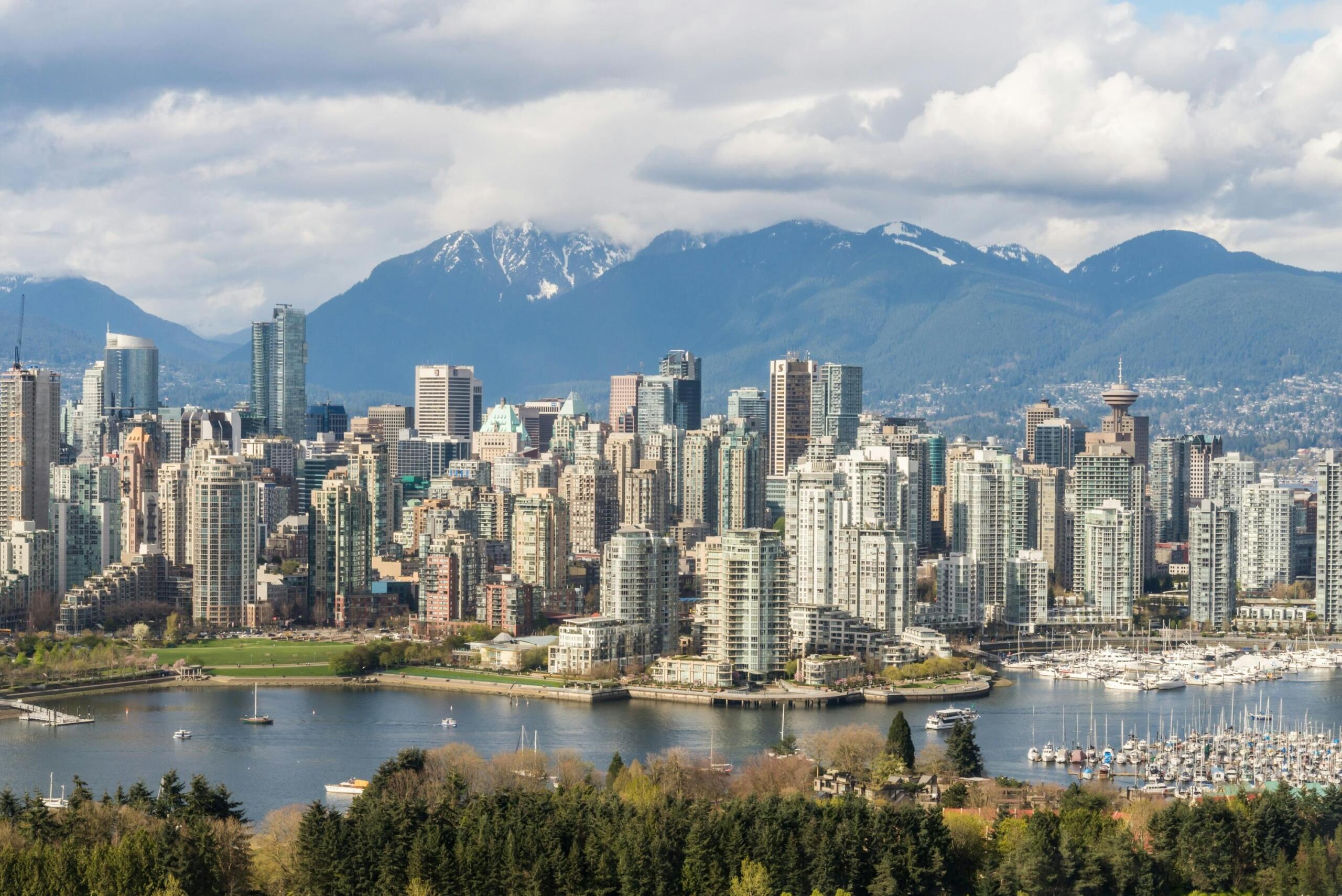 A scenic view of Vancouver Harbour with calm blue water, boats and seaplanes docked along the waterfront, and the downtown skyline rising in the background, framed by distant coastal mountains under a partly cloudy sky.