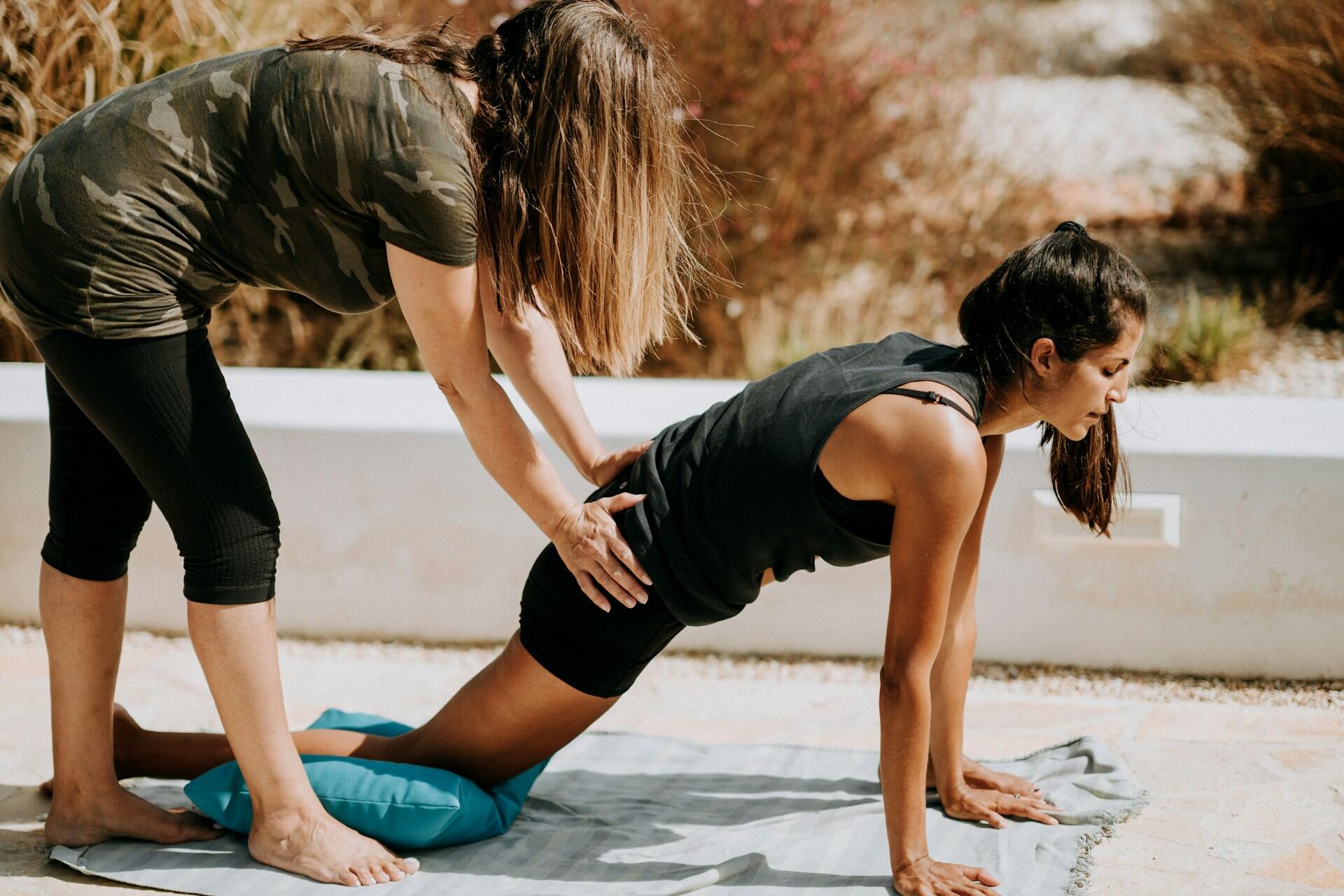 Two women outdoors practicing assisted yoga stretching on a mat near a low white wall and dry foliage.