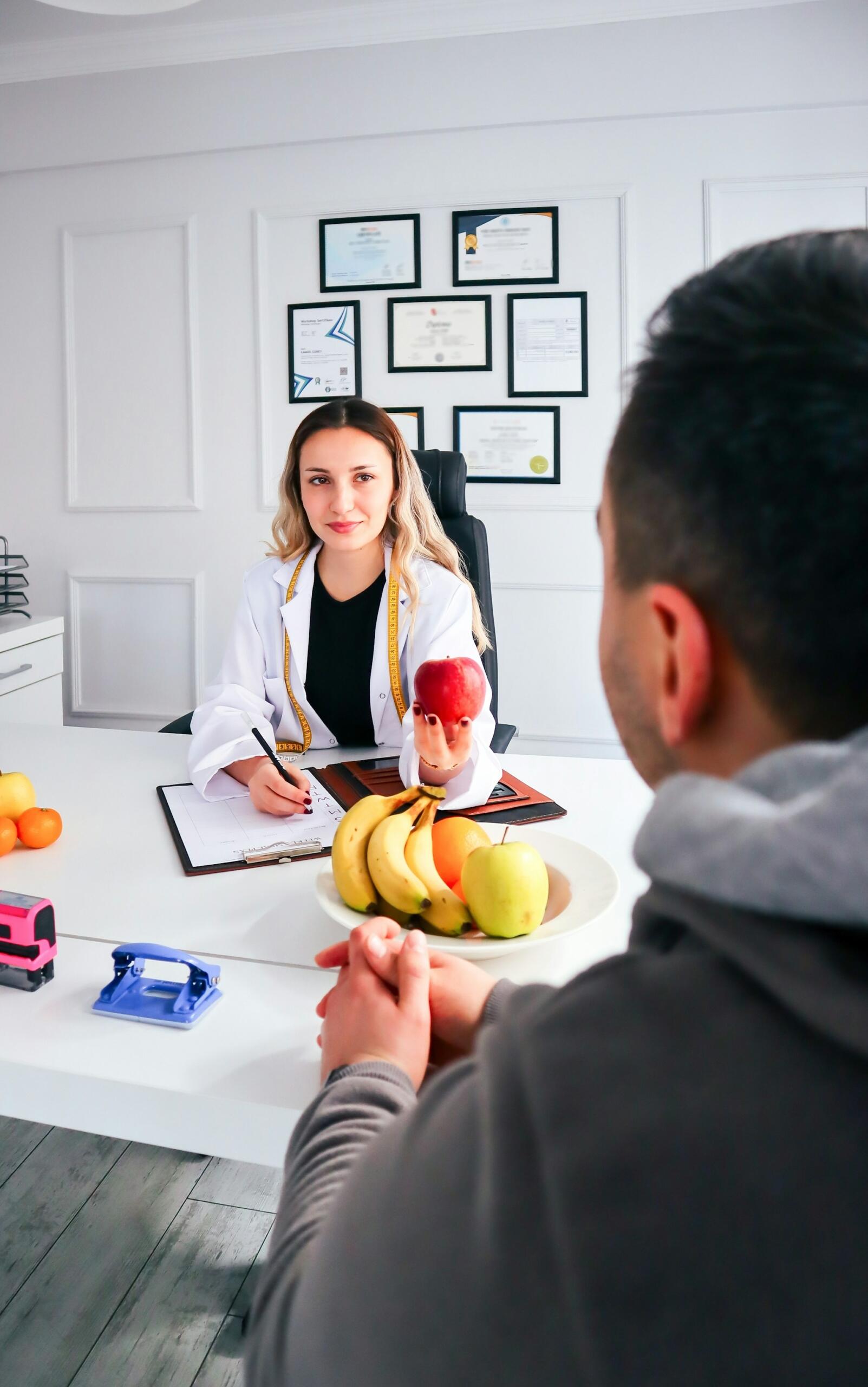 Nutritionist in white coat holding an apple, consulting a patient across a desk with a fruit plate and medical certificates on the wall.