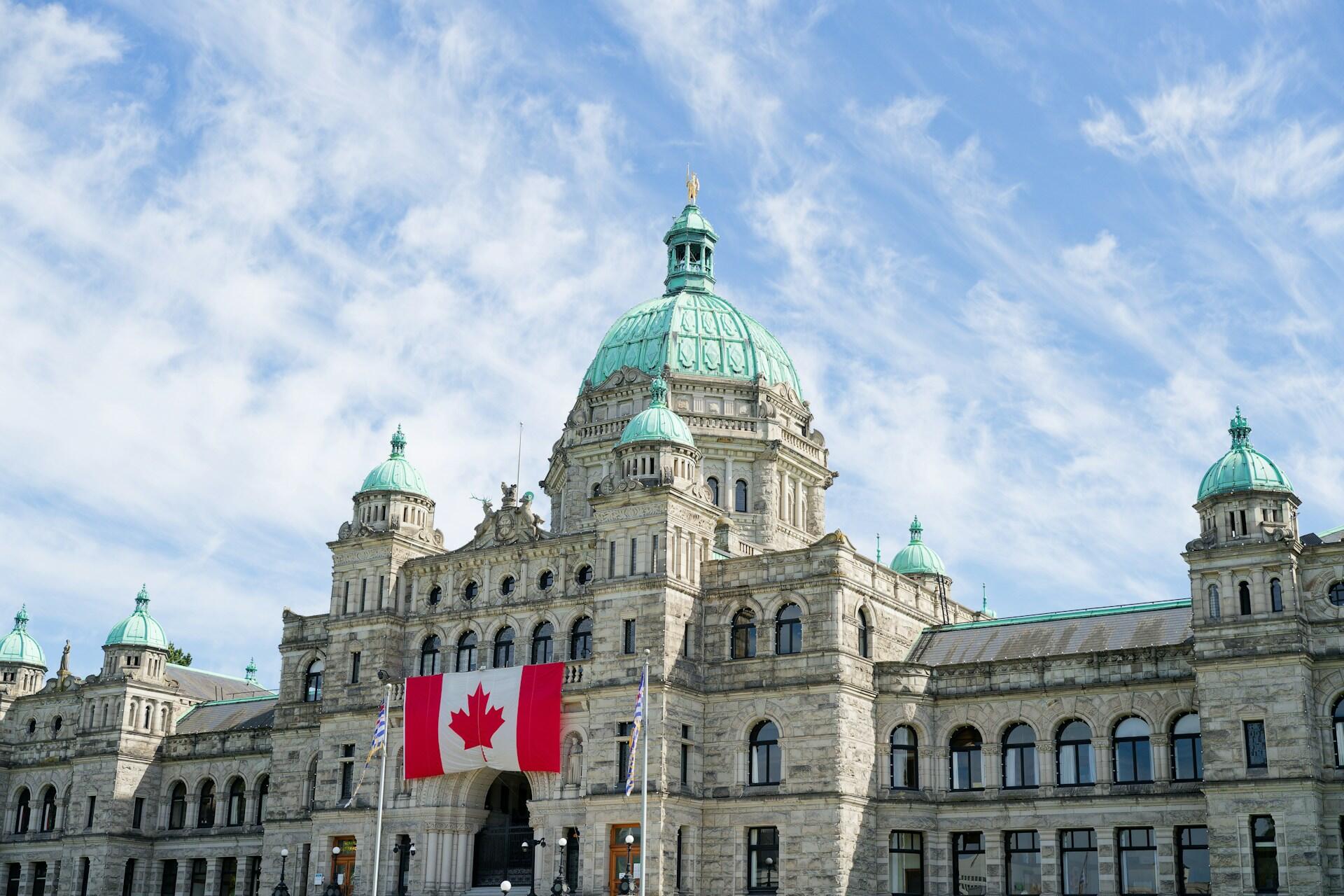 The British Columbia Parliament Buildings with green domes and a large Canadian flag hanging in front, under a partly cloudy sky.