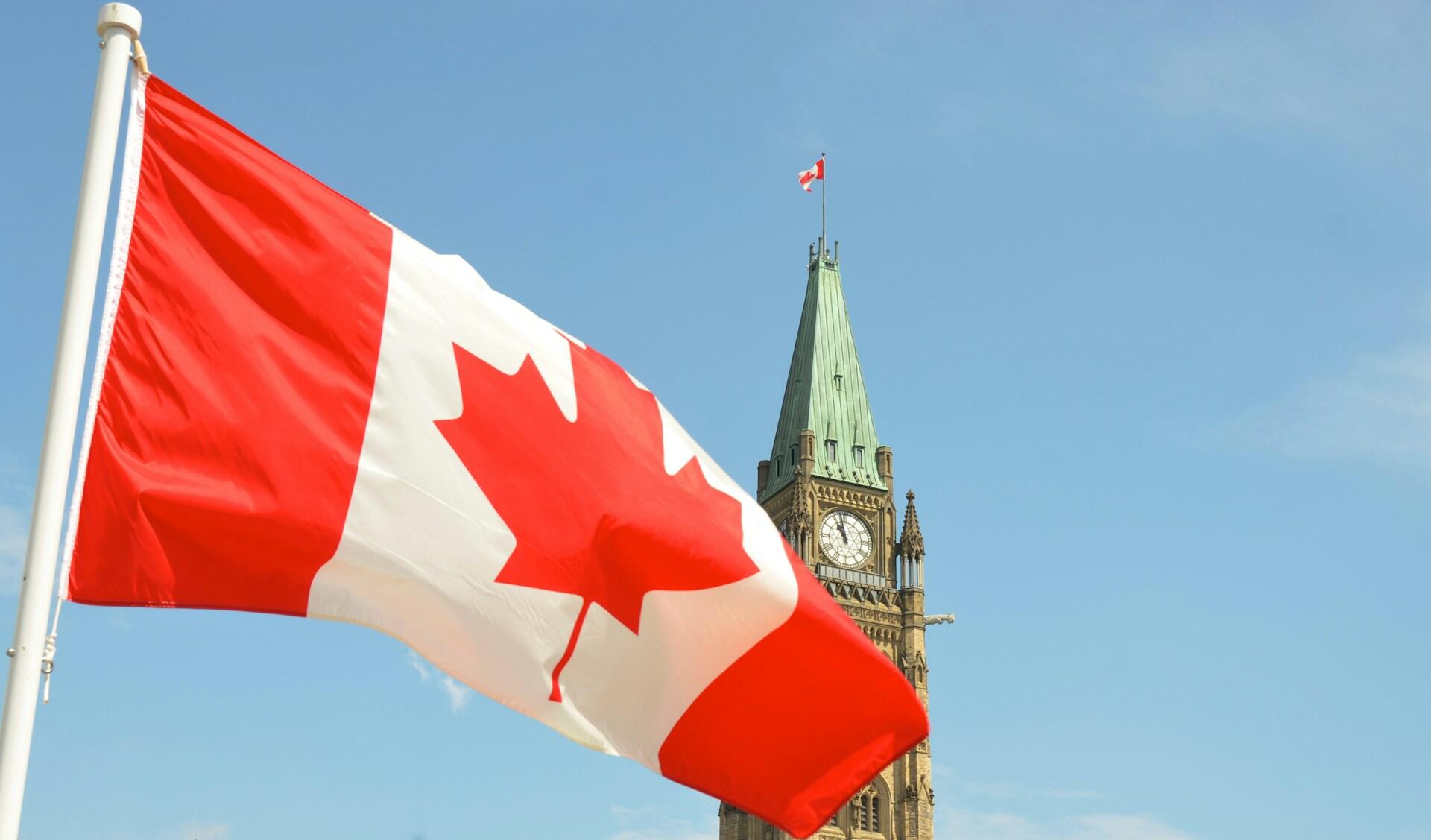 Canadian flag waves in the foreground with the Peace Tower of Parliament Hill and a clear blue sky in the background.