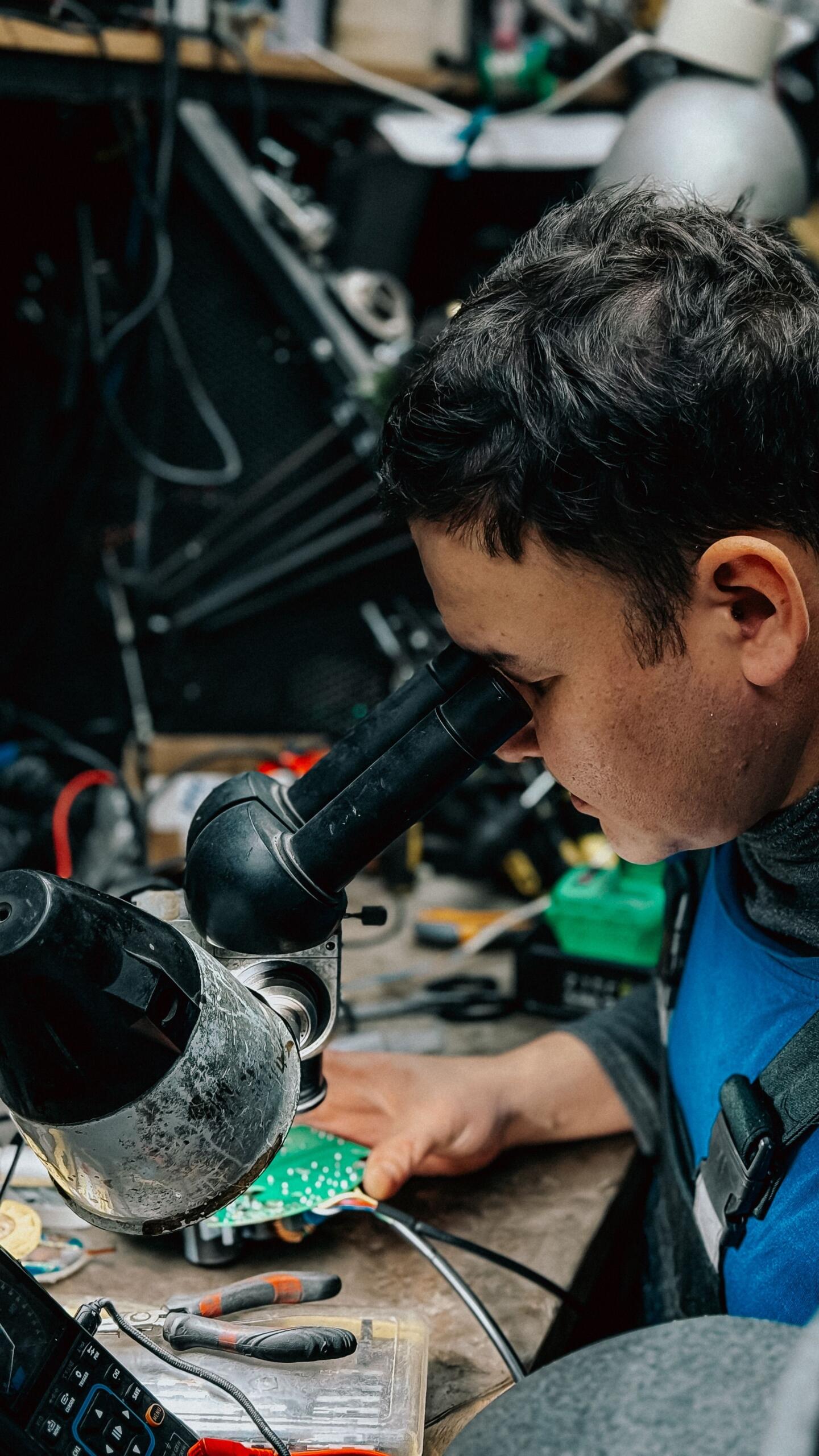 A man peers through a microscope while closely examining a circuit board in a laboratory or technology workspace.